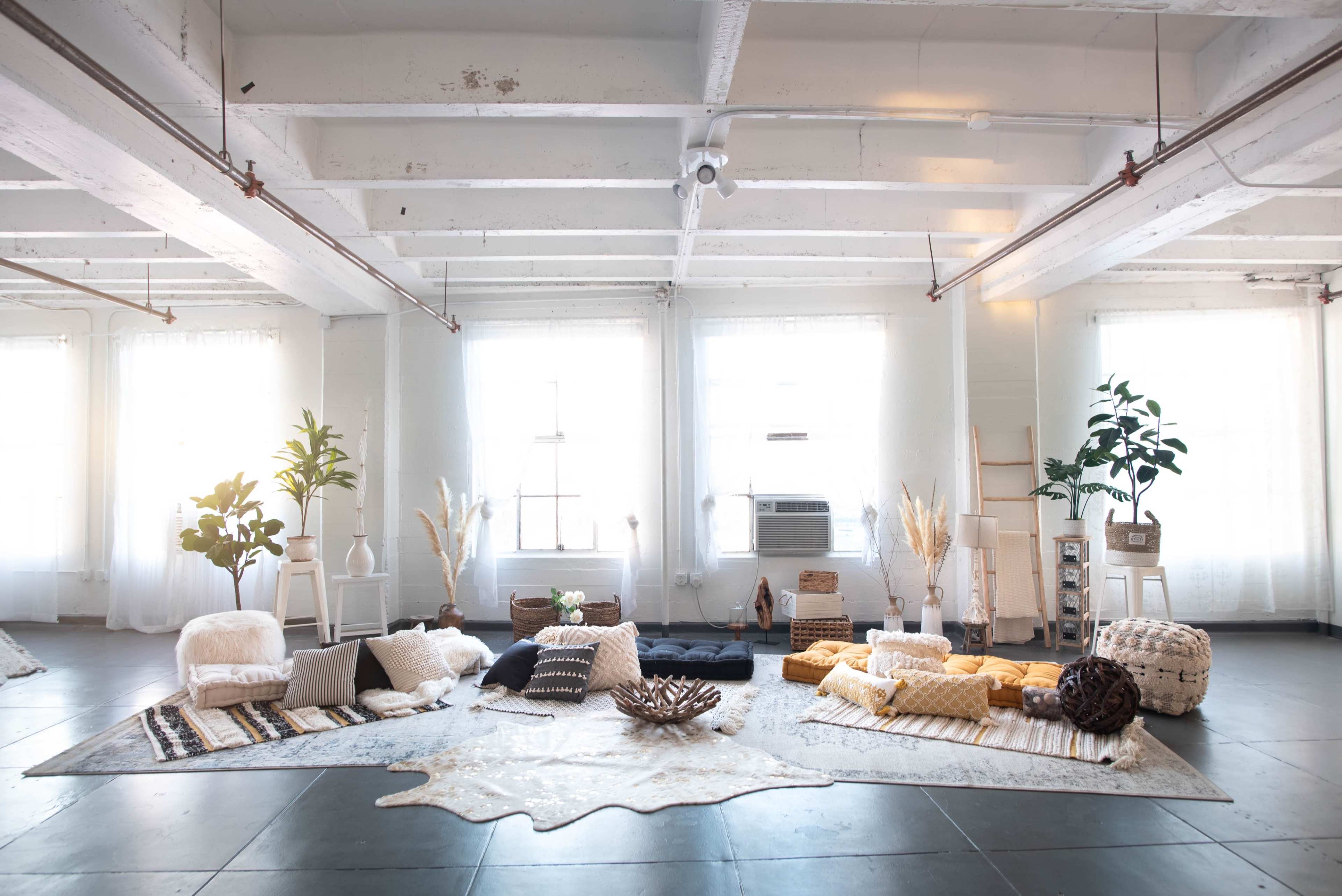 A spacious, bright room features various cushions, plants, and decorative elements arranged on a textured rug.