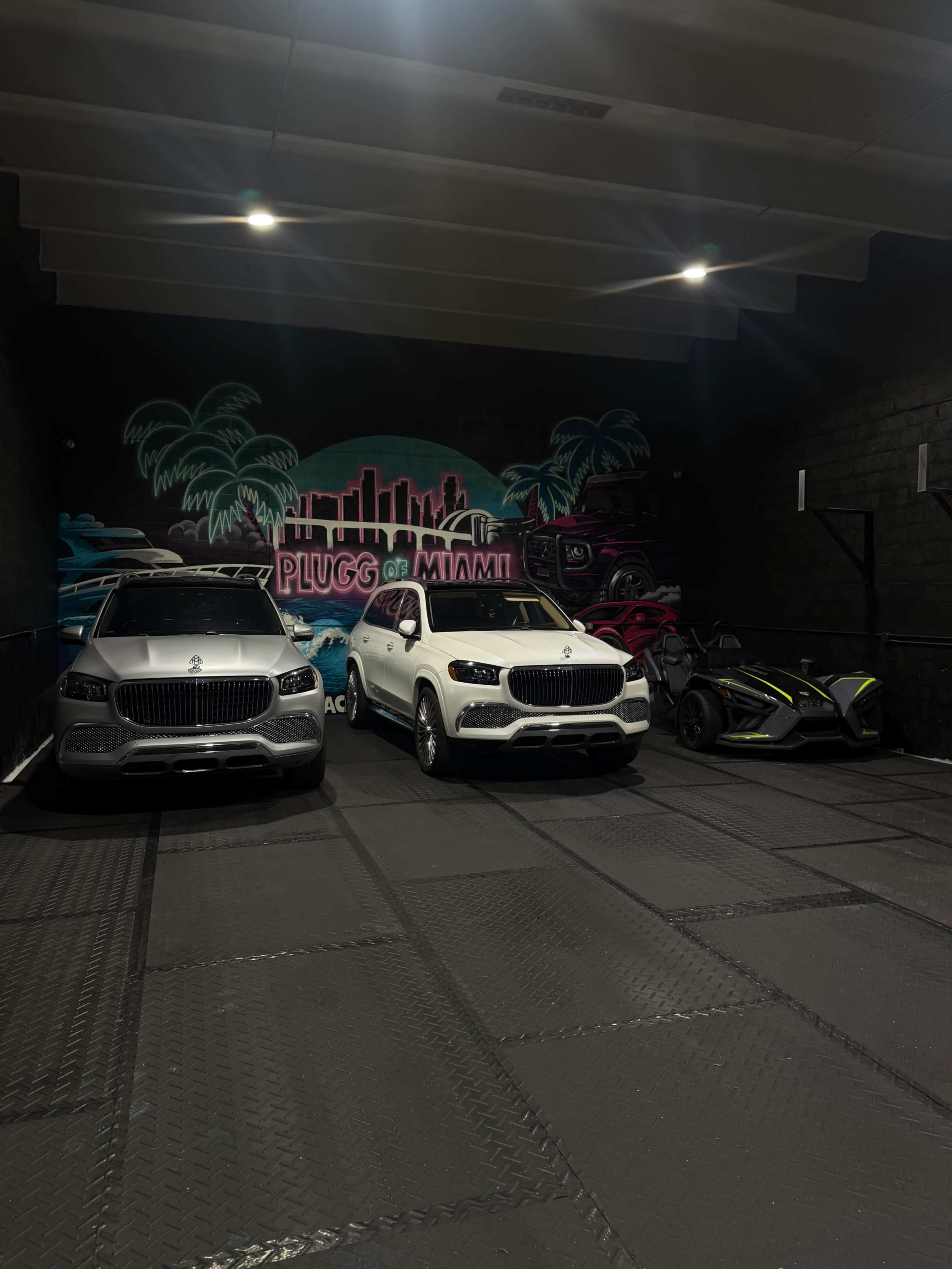 The image shows a garage with three vehicles: two luxury SUVs and a sports car, against a colorful mural that reads "PLUGG OF MIAMI."