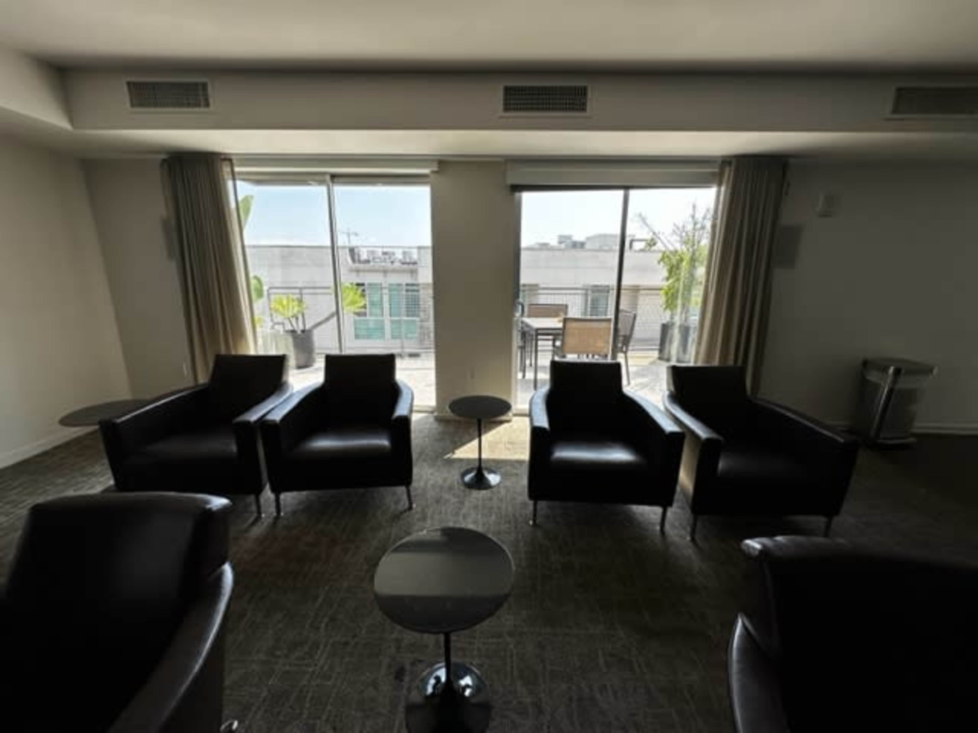 A modern lounge area features four leather chairs arranged around small tables, with large windows providing a view of an outdoor terrace.