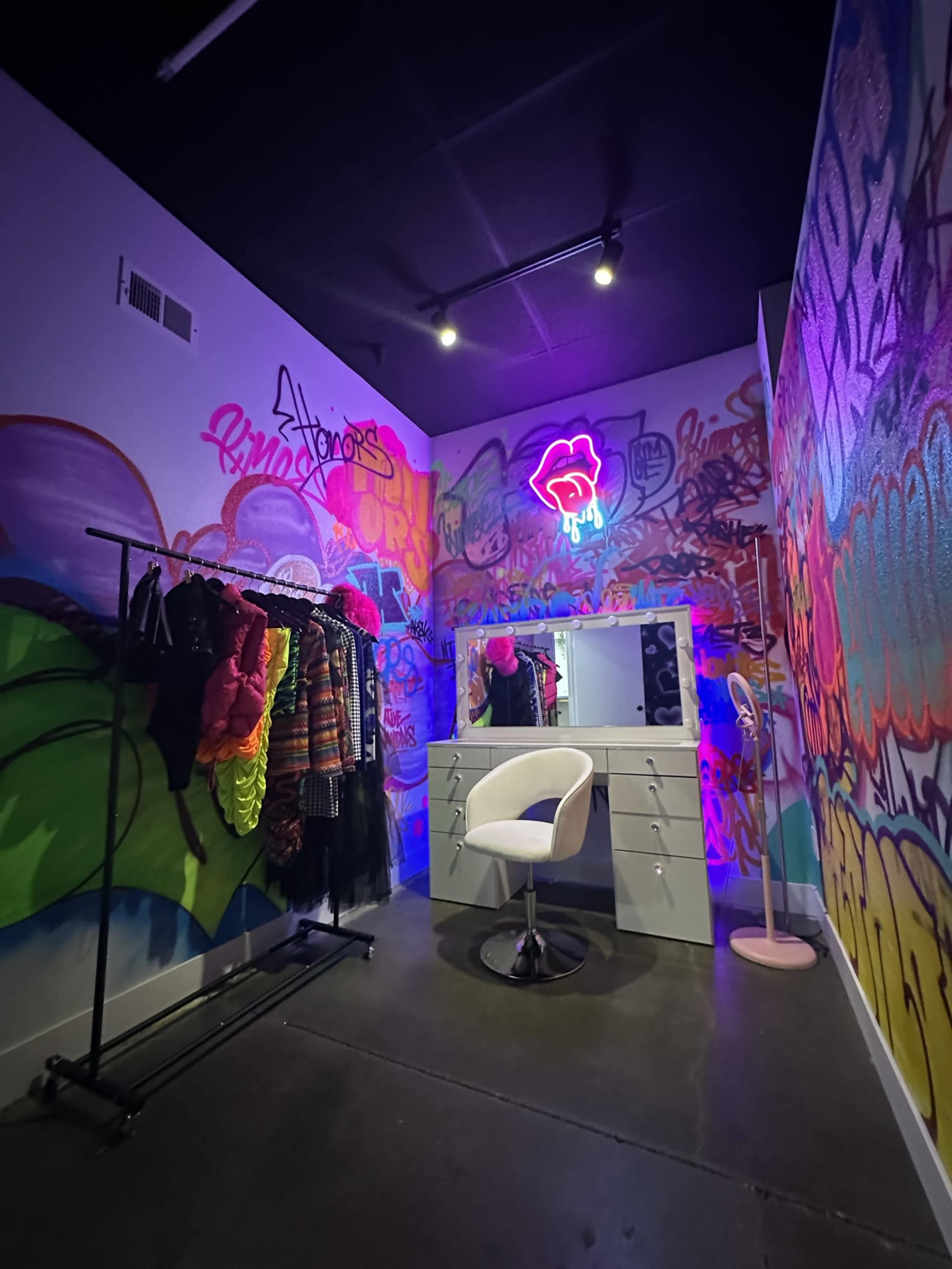 A brightly colored room with graffiti-covered walls, featuring a clothing rack with various outfits, a white vanity with a mirror, and a neon sign.