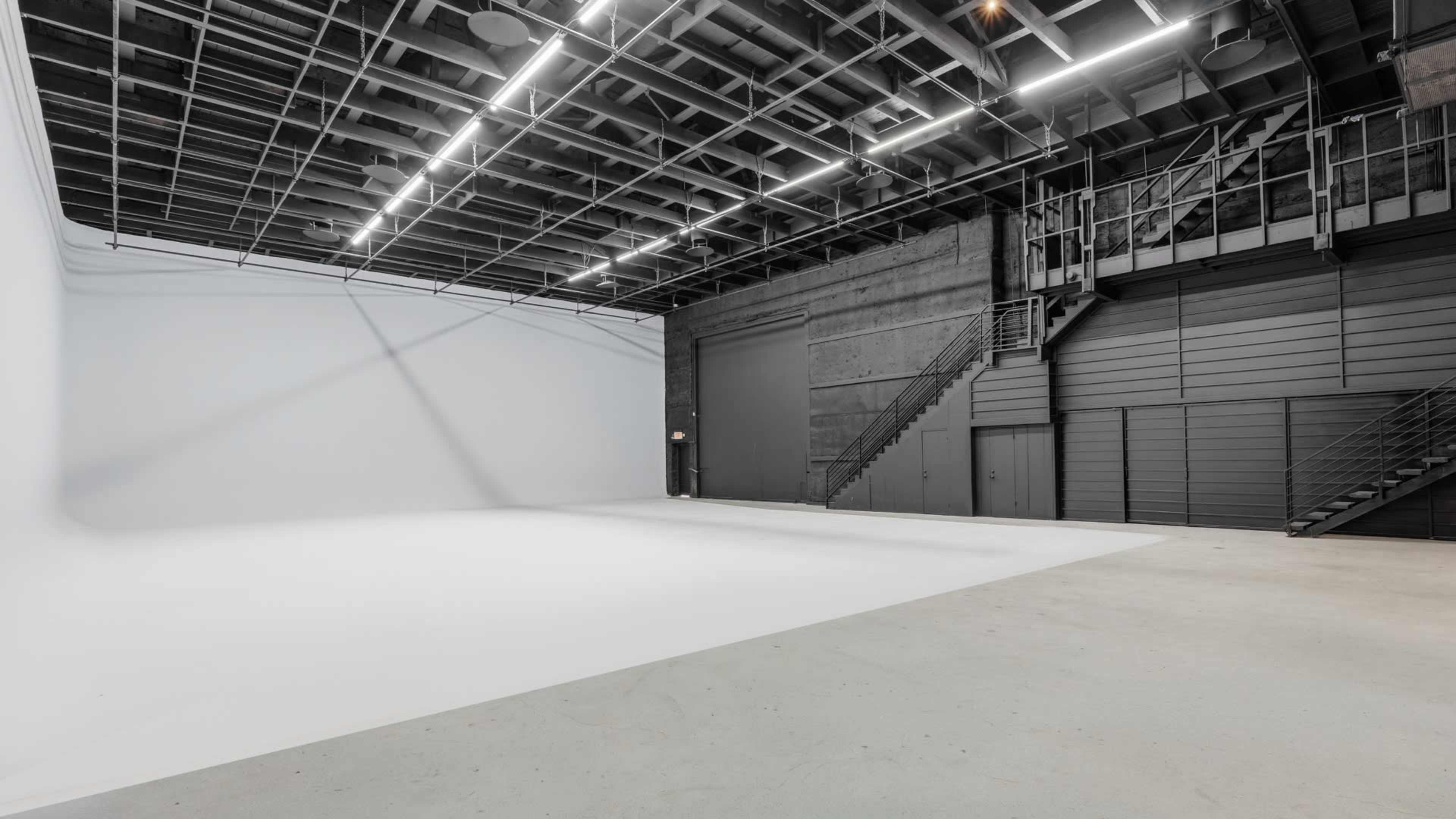 The image shows a large, empty studio space with a smooth white backdrop and a high ceiling, featuring metal stairs and industrial-style walls.