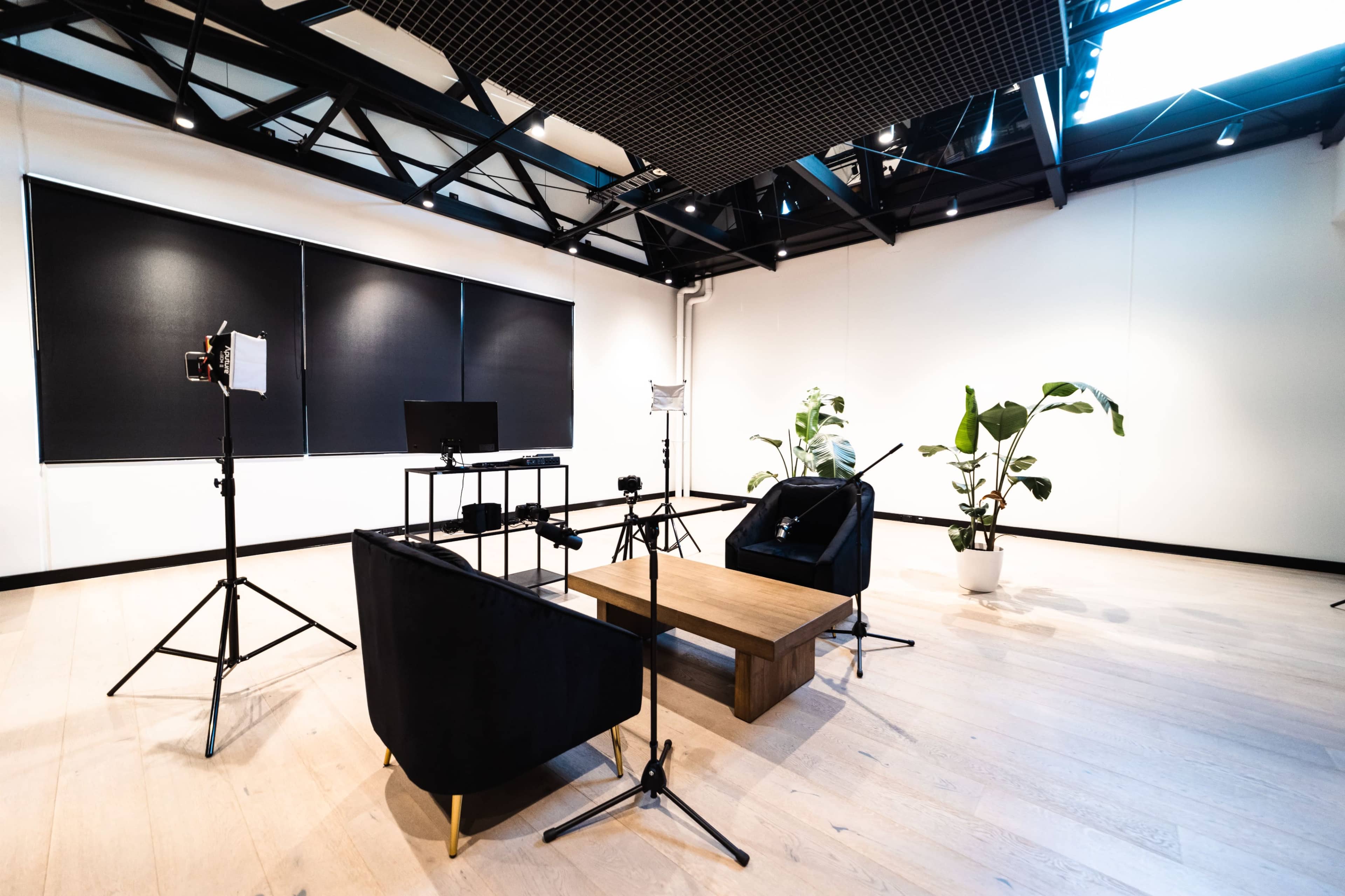 A modern studio setup with two chairs, a coffee table, and lighting equipment arranged around a potted plant.