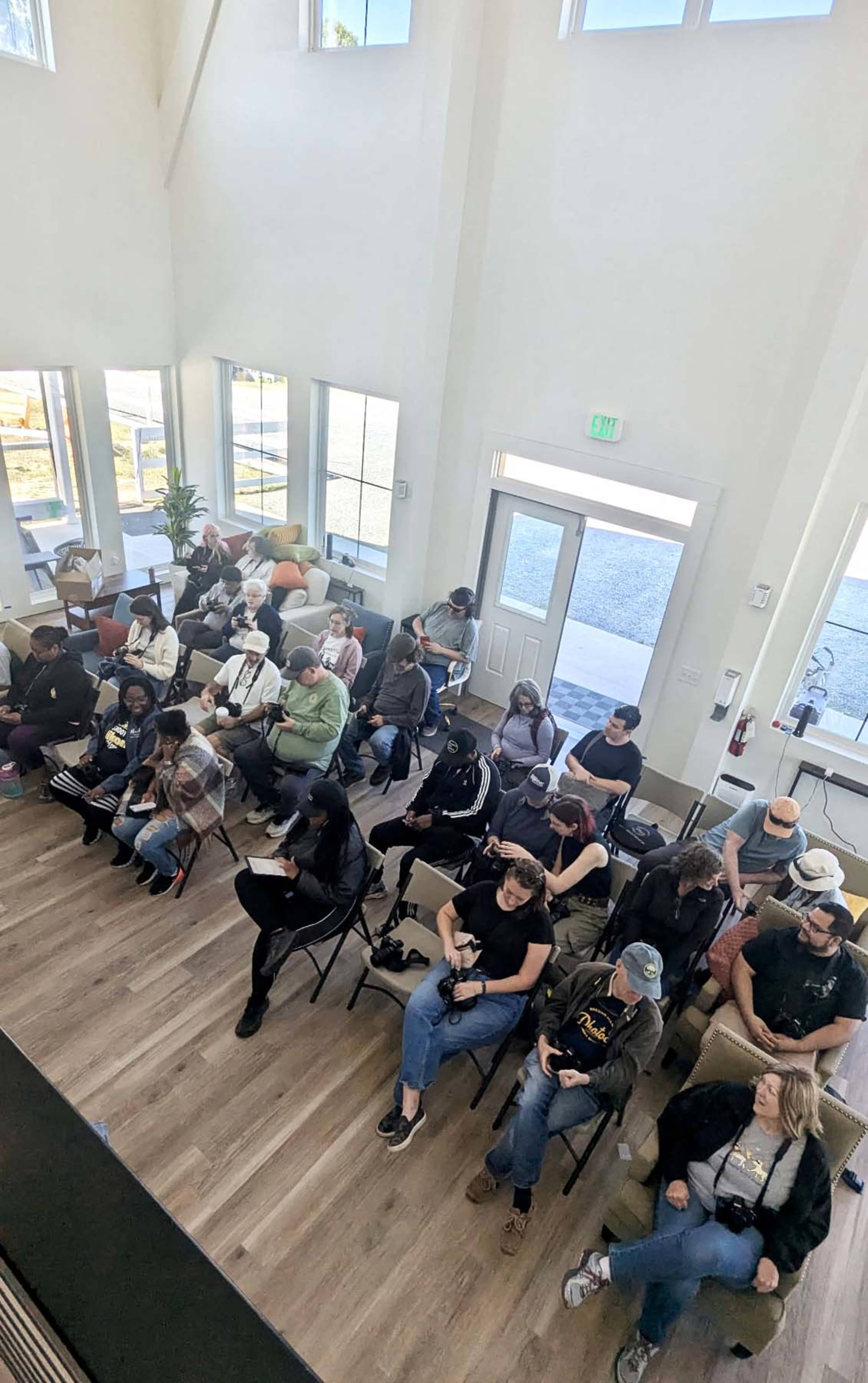 A group of people sits in chairs organized in rows inside a well-lit room with large windows.