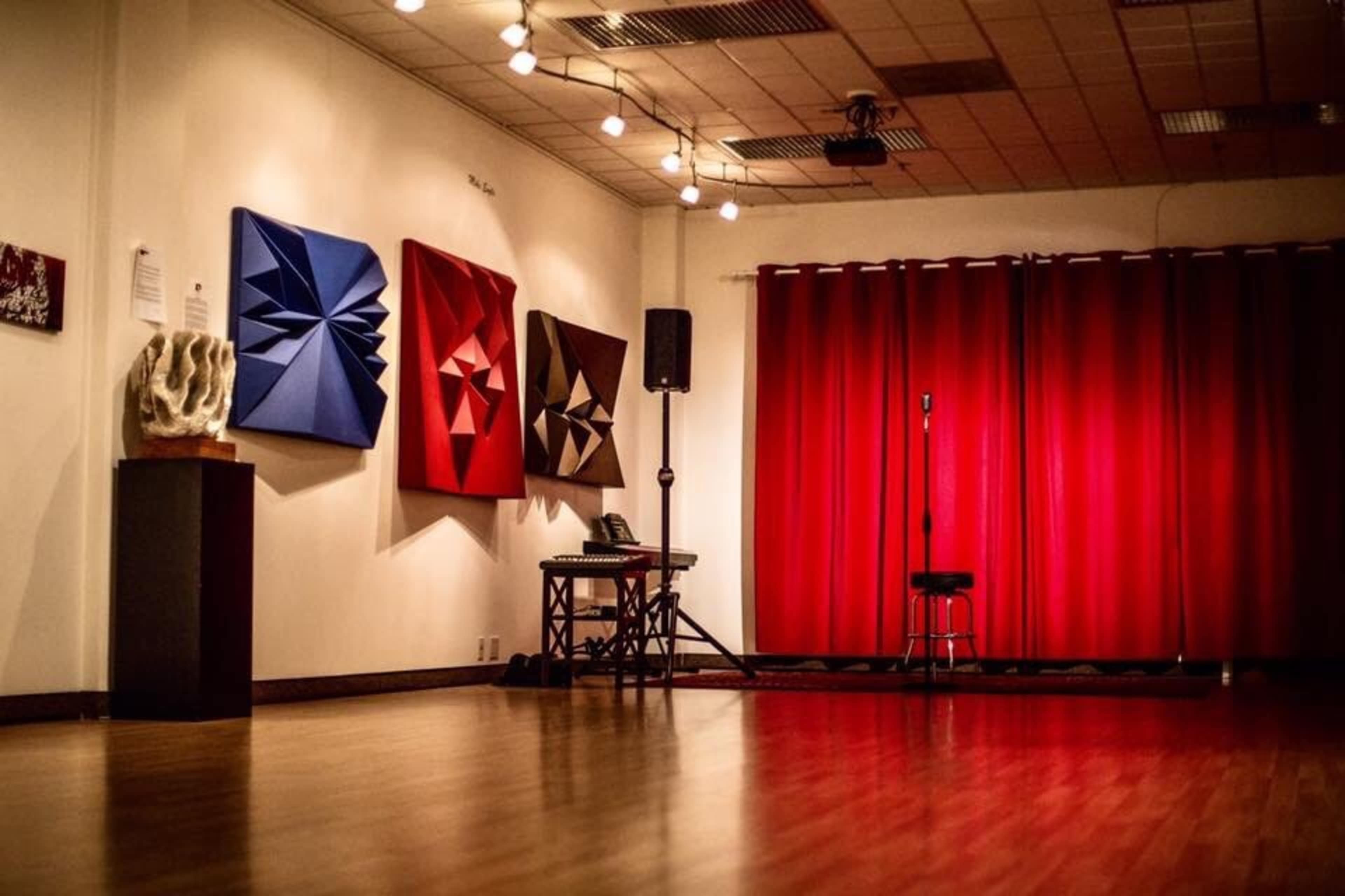 The image depicts an art gallery with three colorful geometric wall sculptures, a microphone stand, and a red curtain in the background.