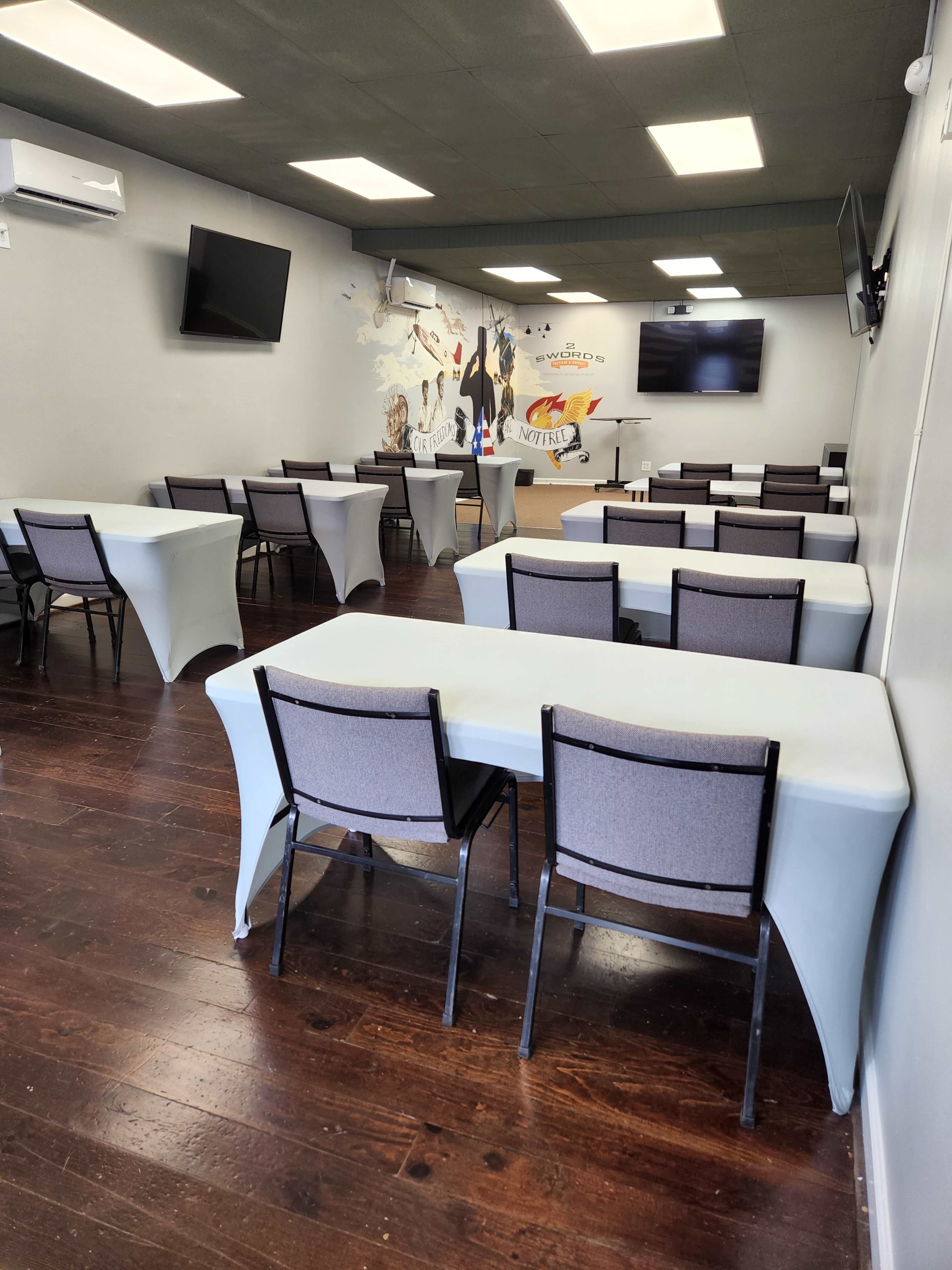 A spacious room set up with six rows of table-and-chair arrangements, featuring a mural on one wall and television screens mounted on the opposite walls.