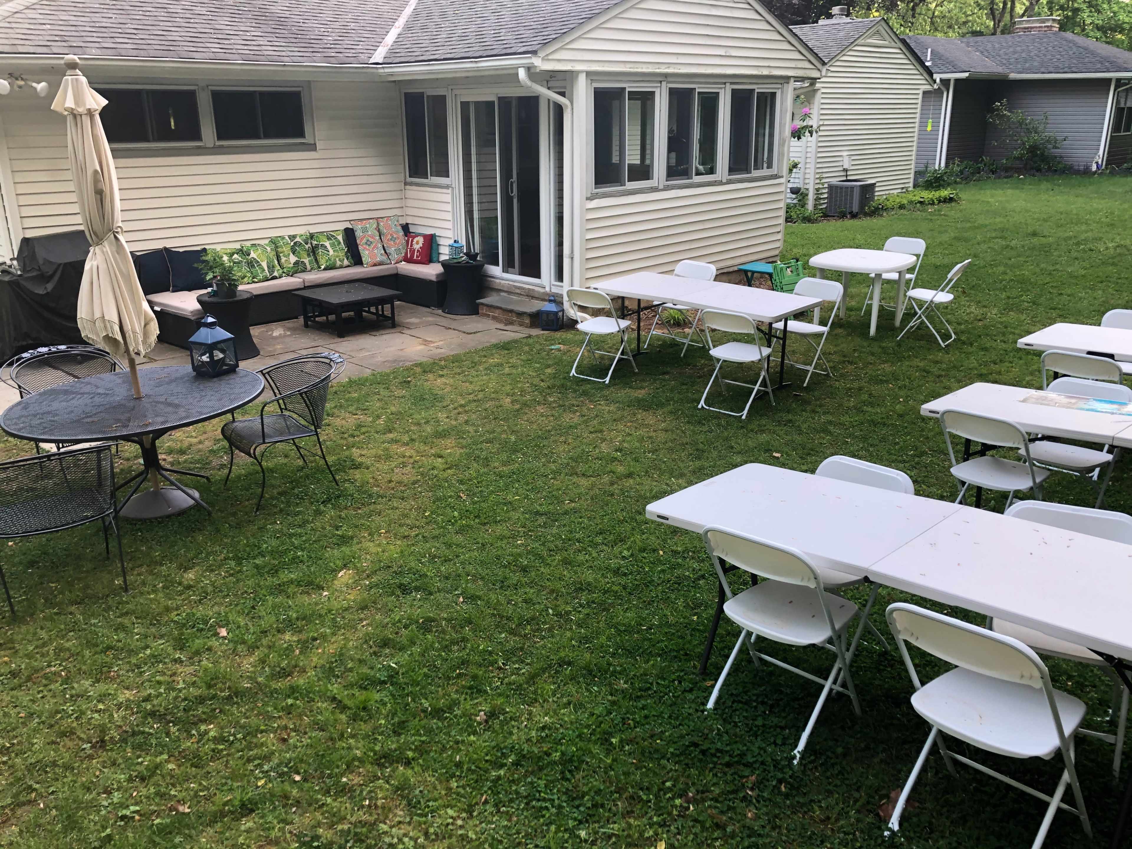A grassy backyard features several empty folding tables and chairs arranged for an outdoor gathering, with a house visible in the background.