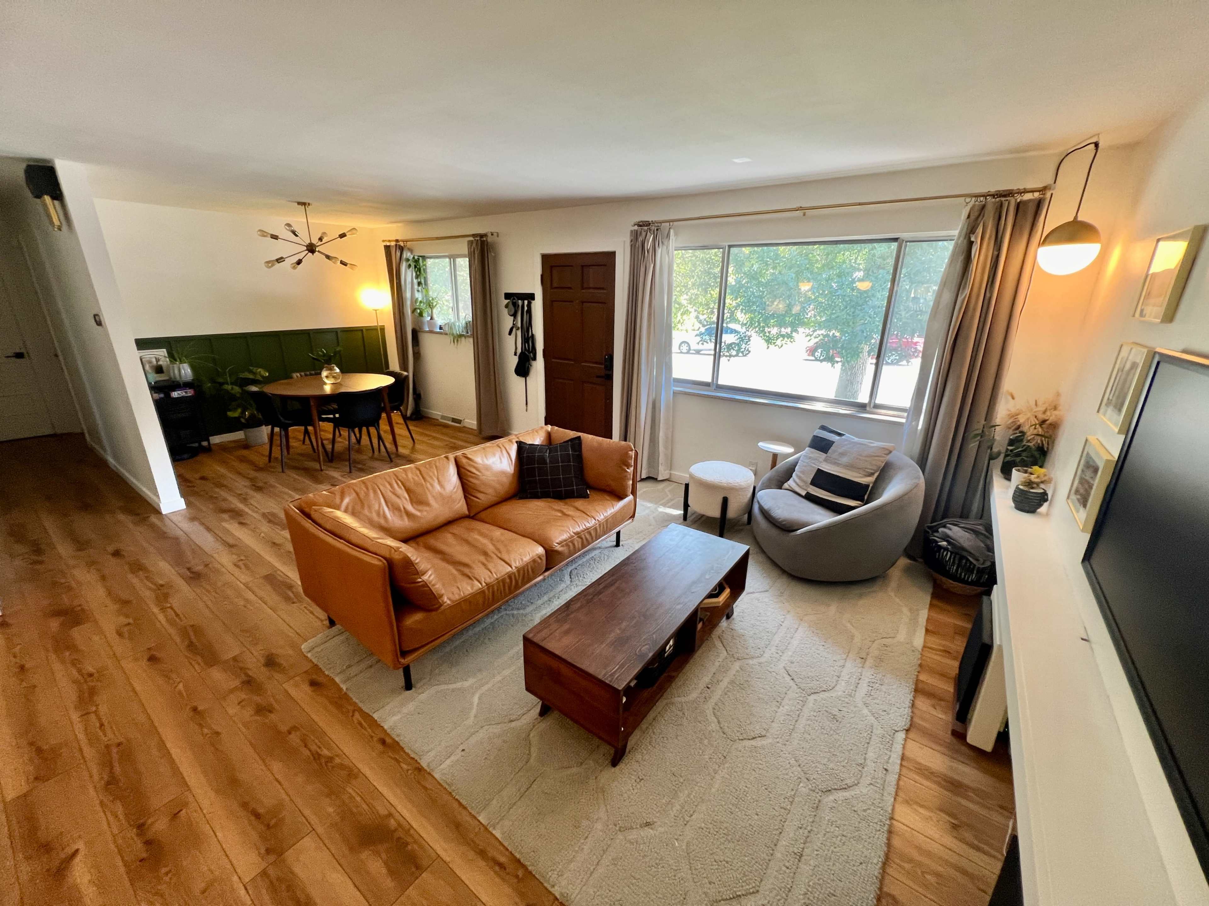 The image shows a living room with a brown leather sofa, a circular chair, a wooden coffee table, and a large window letting in natural light.