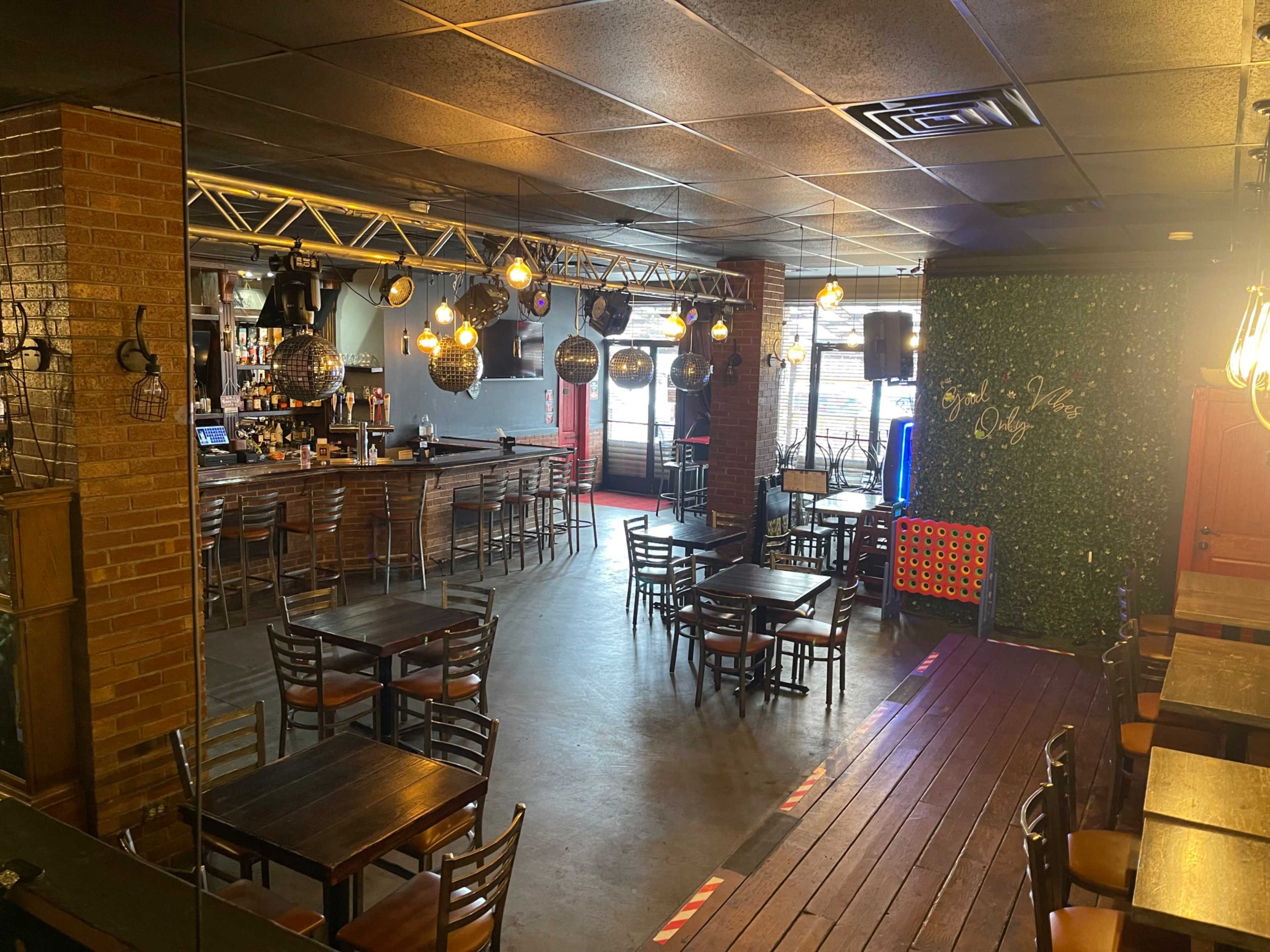 The image shows an interior of a bar with tables and chairs arranged in a spacious layout, a wooden bar counter, and decorative lighting.