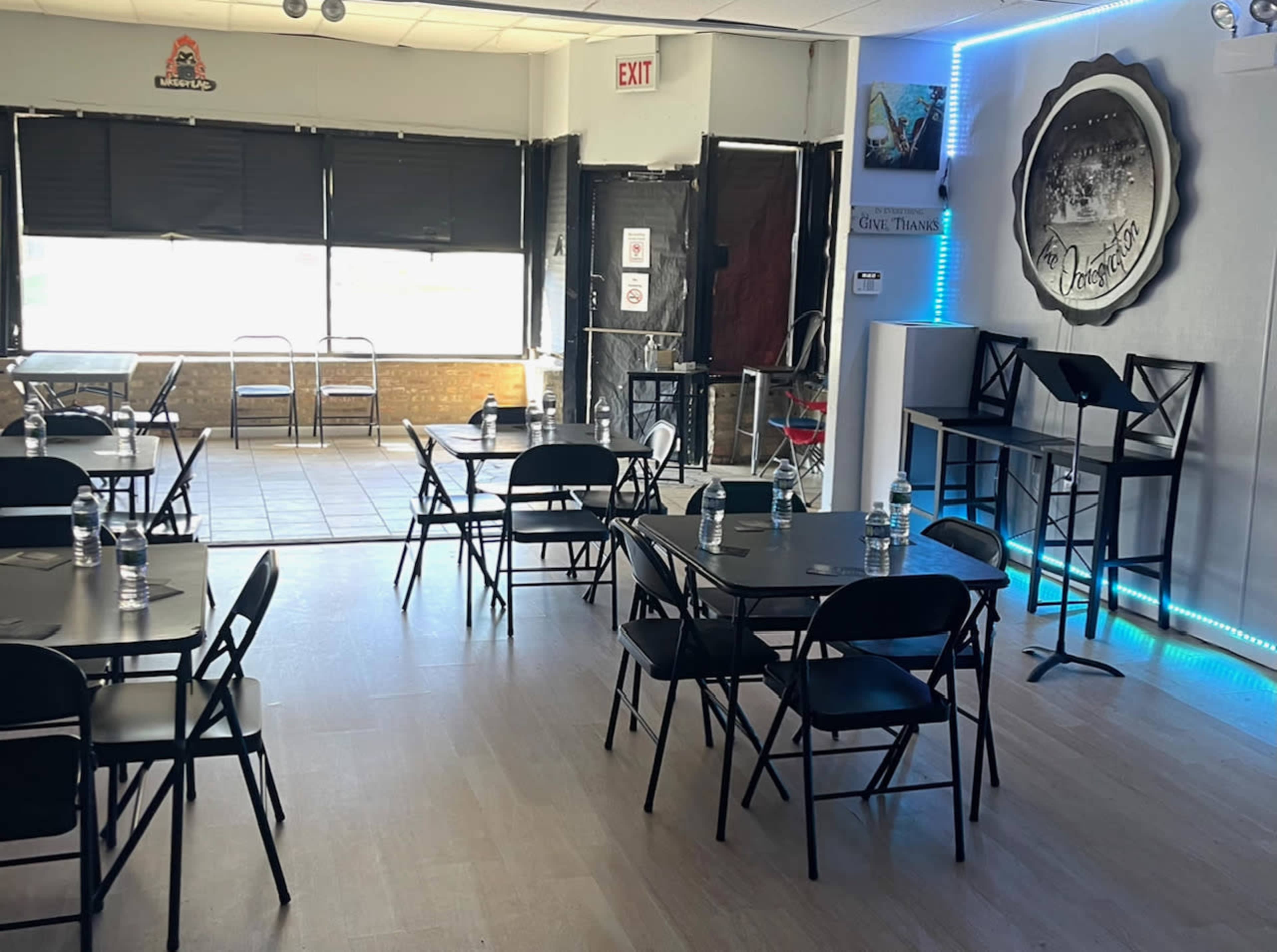 An empty indoor space with several black folding tables and chairs, a podium, and large windows letting in natural light.