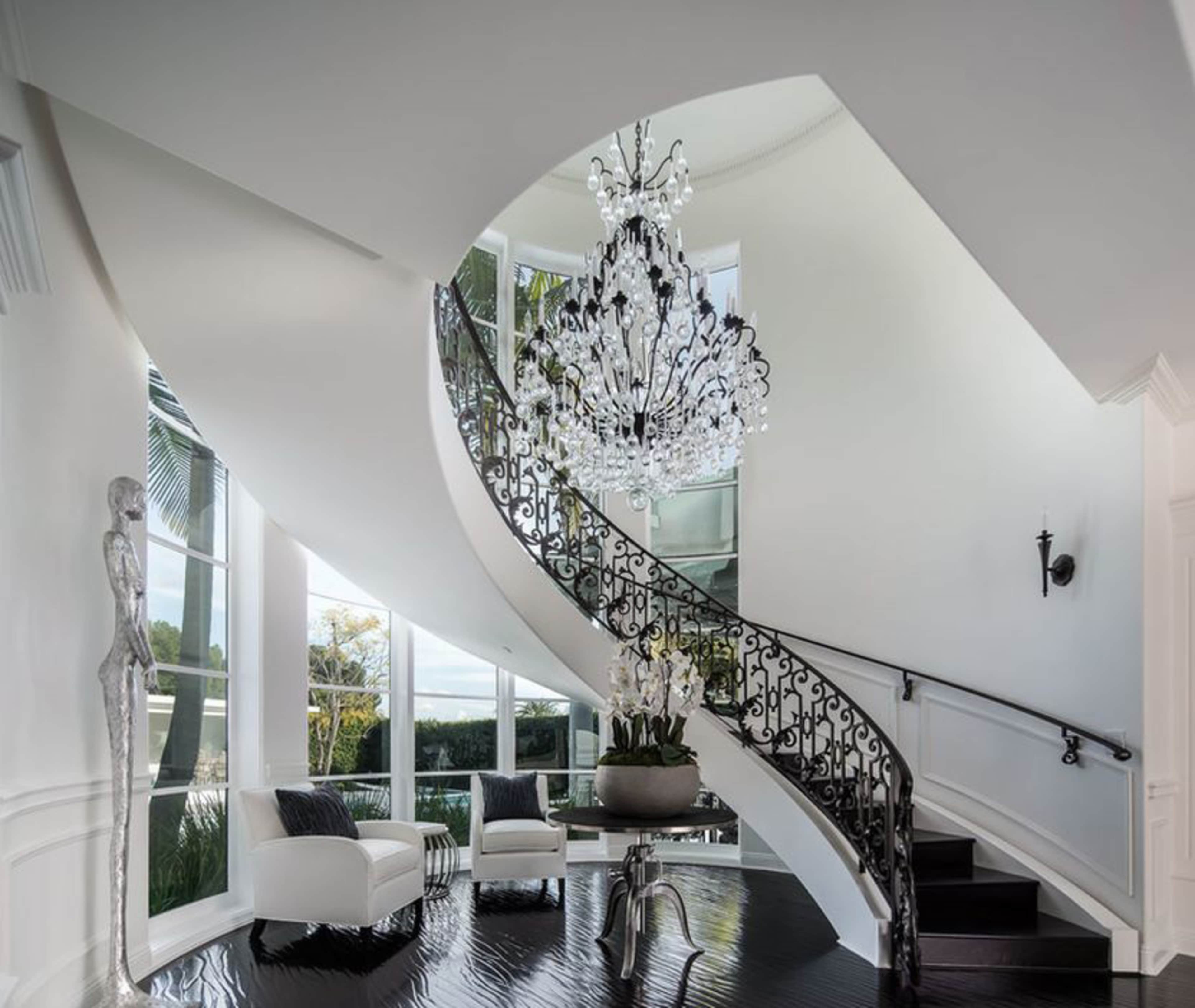 A grand staircase with a wrought iron railing features a large chandelier and is flanked by modern seating in a well-lit foyer.