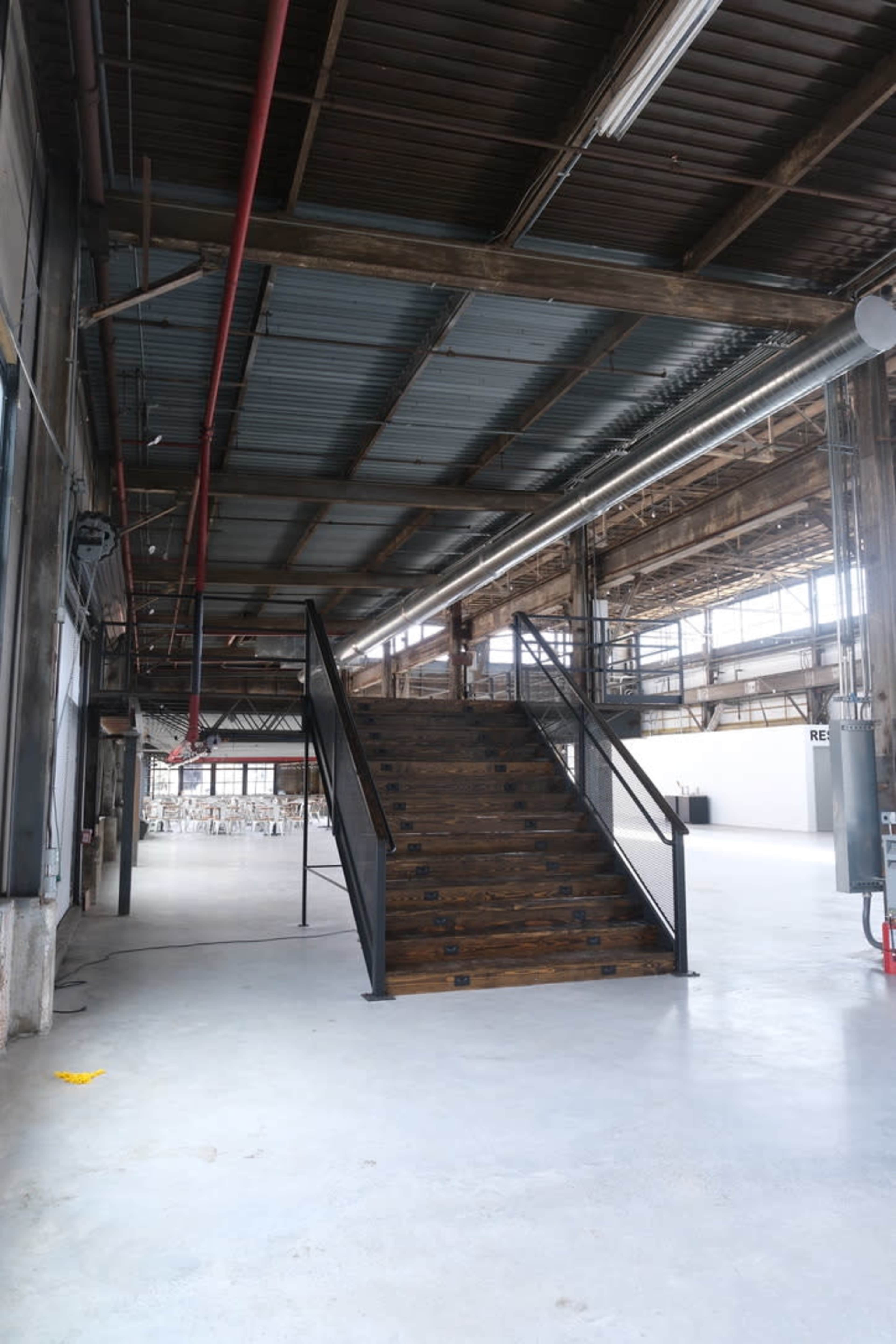 The image shows a set of wooden stairs with a metal railing in an industrial-style building with exposed beams and a concrete floor.