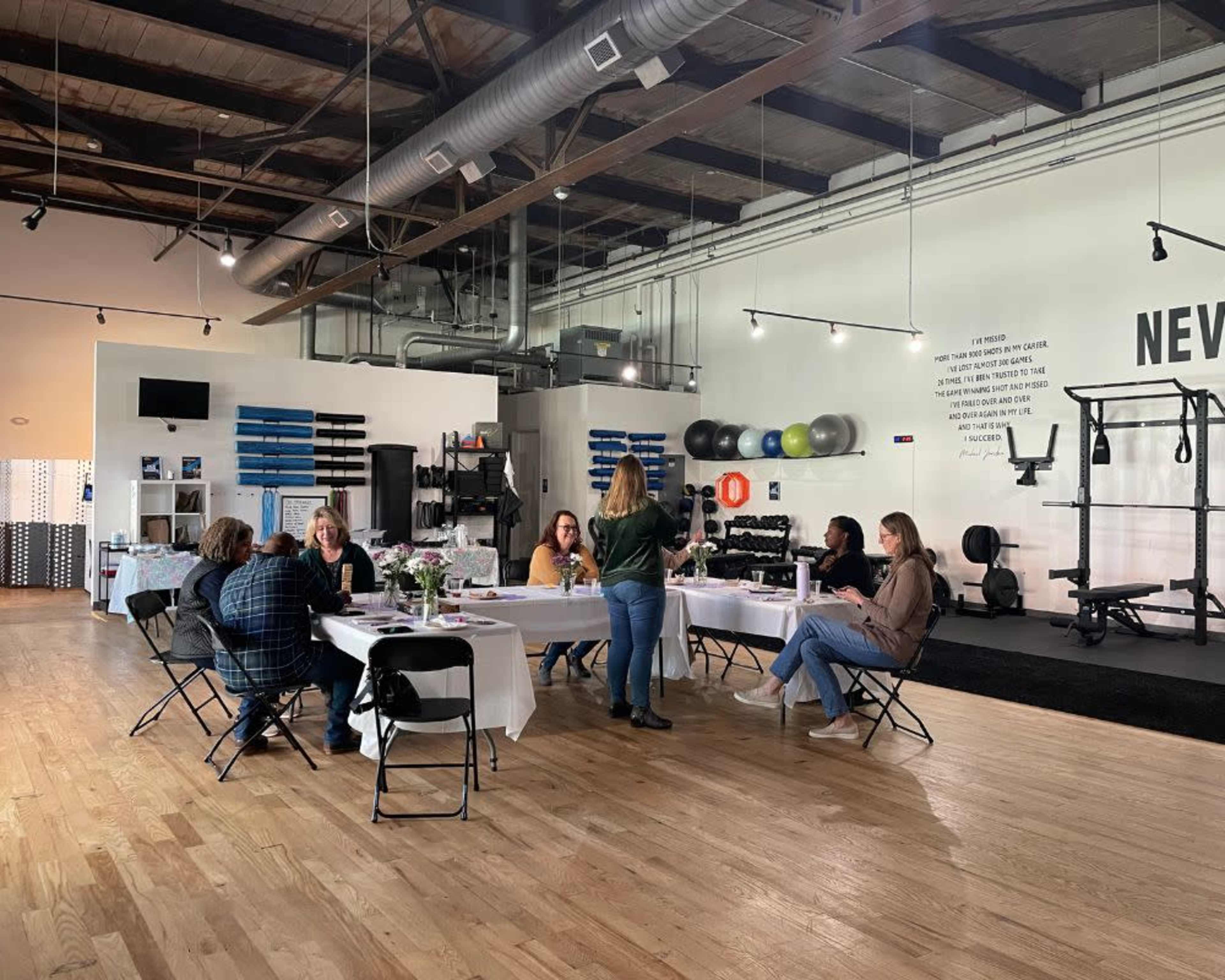A group of people is seated around tables in a spacious room equipped with fitness equipment and decorations on the walls.