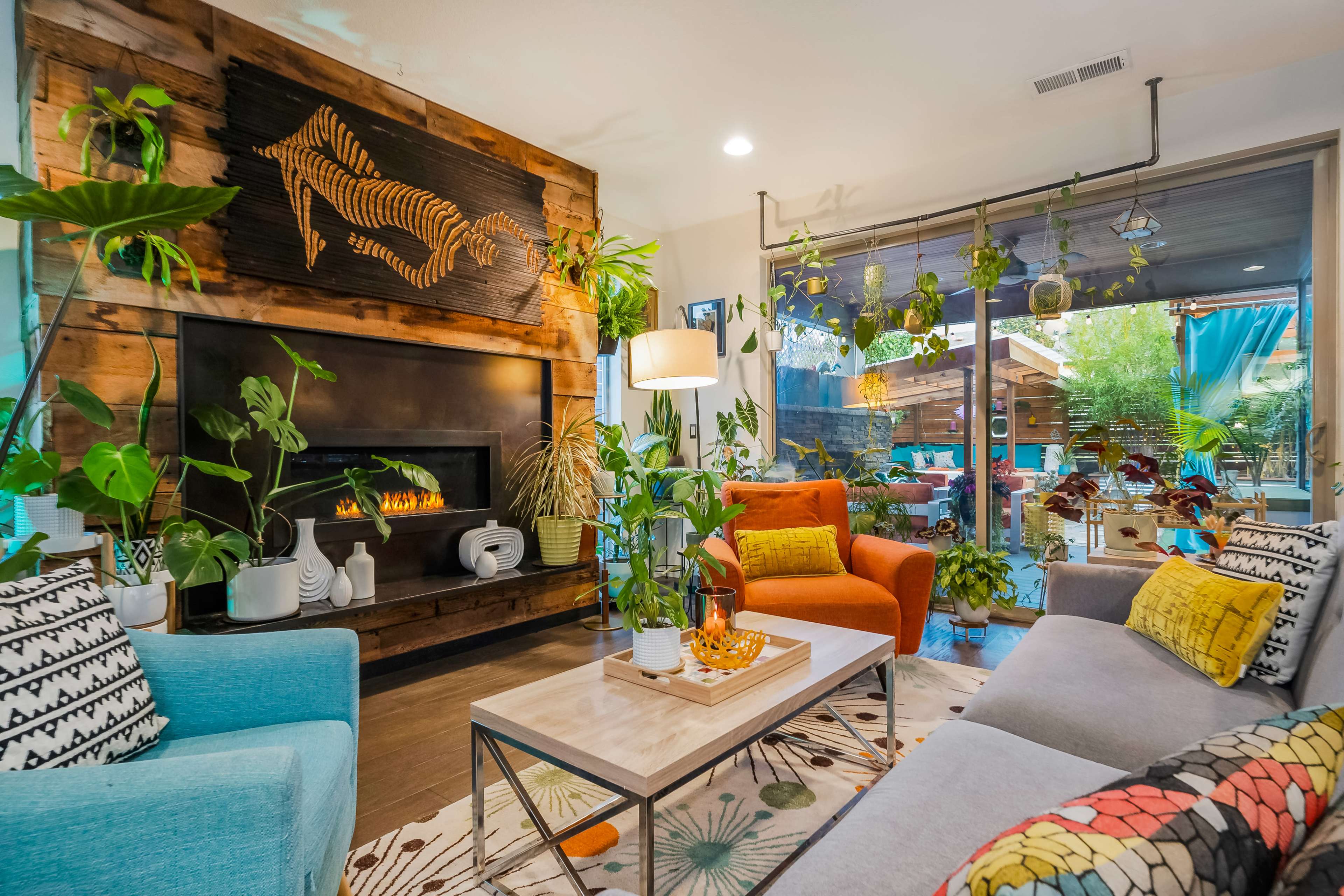 The scene depicts a cozy living room with a fireplace, plants, and colorful furniture, including a blue sofa and an orange chair, surrounding a coffee table on a patterned rug.