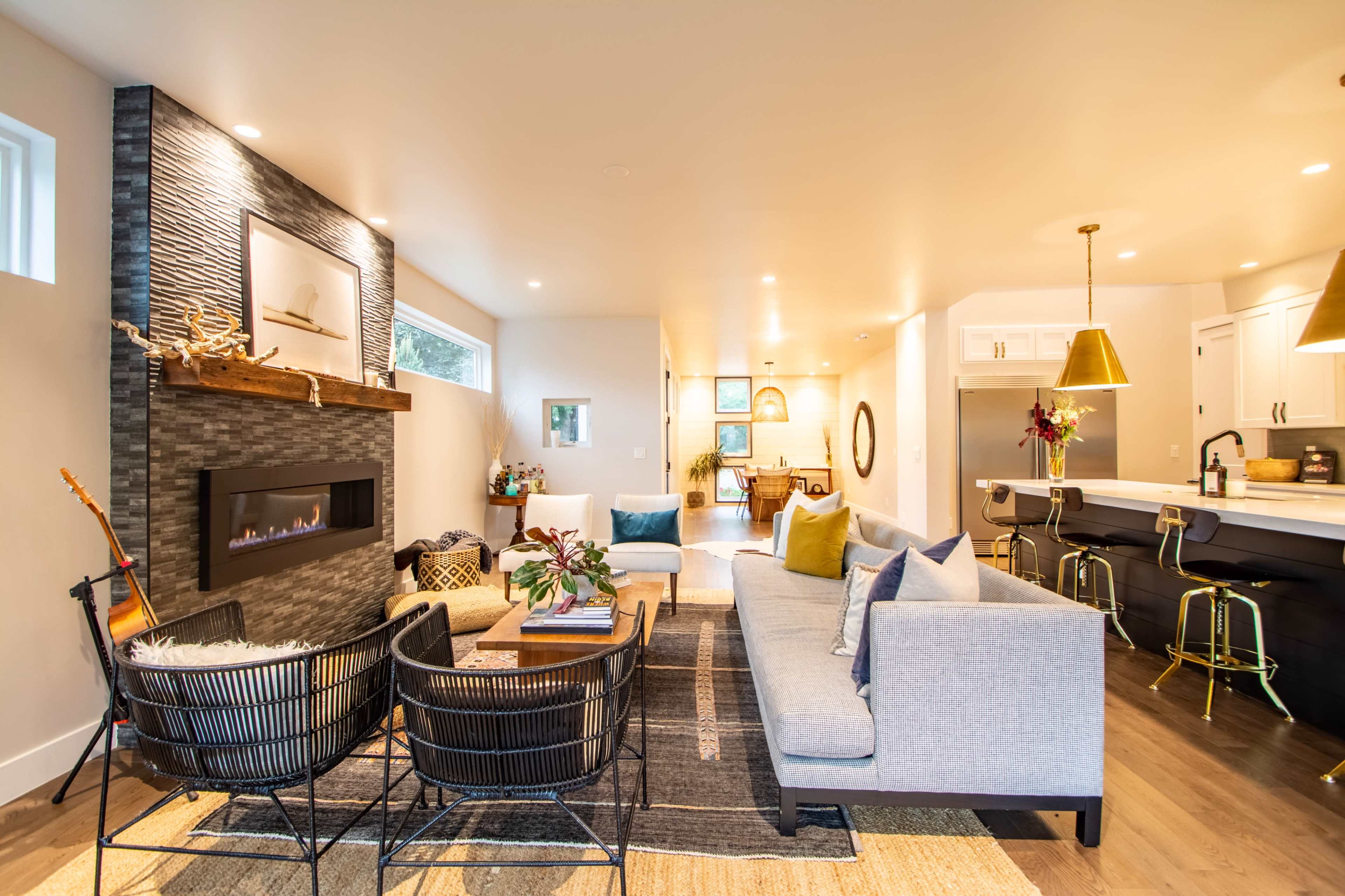 The image shows a modern living room featuring a gray sofa, two black wire chairs, a wall-mounted fireplace, and a view into a light-filled kitchen area.