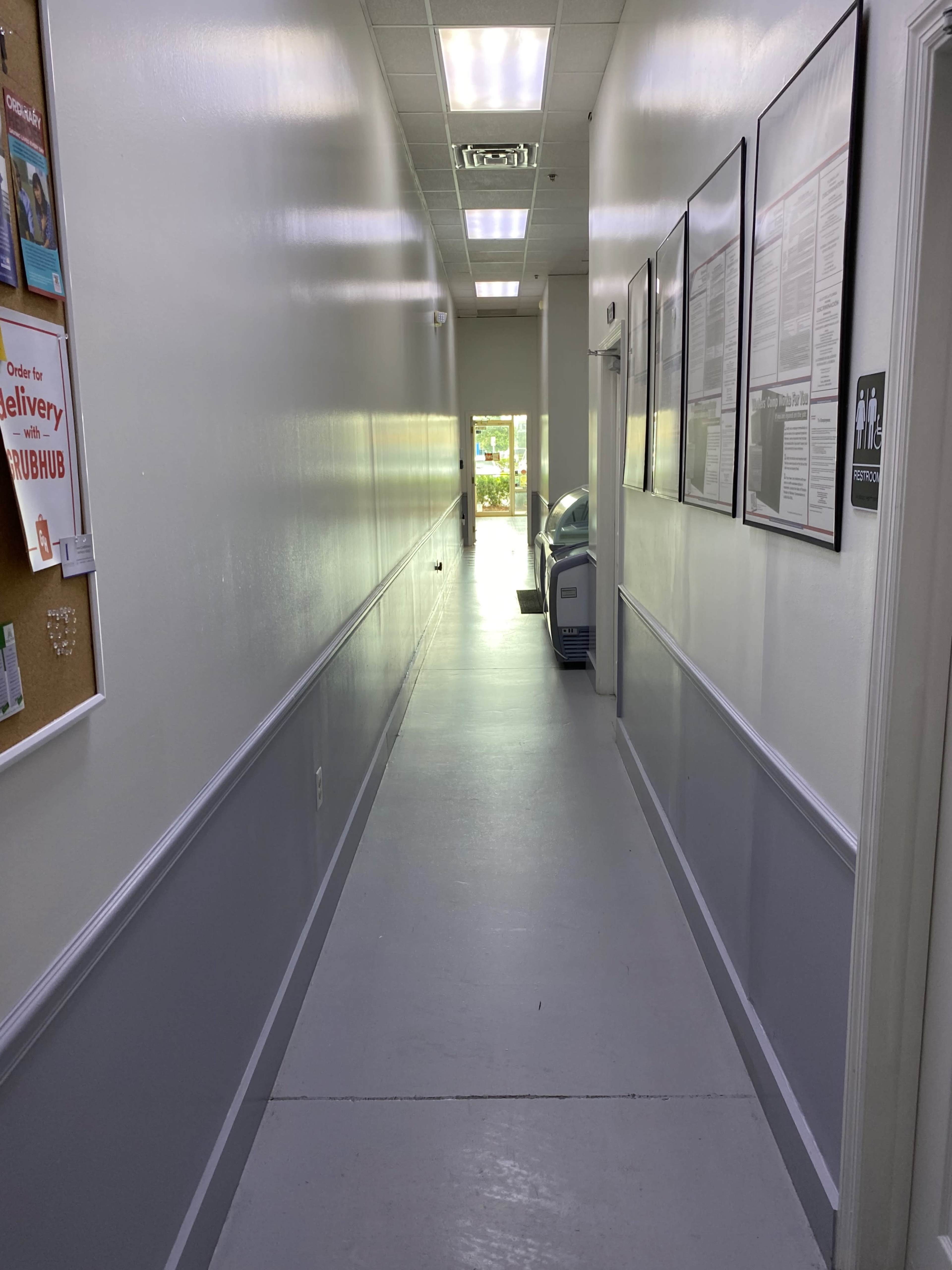 The image shows a narrow, well-lit hallway with white walls, a gray floor, and a door at the end leading outside.