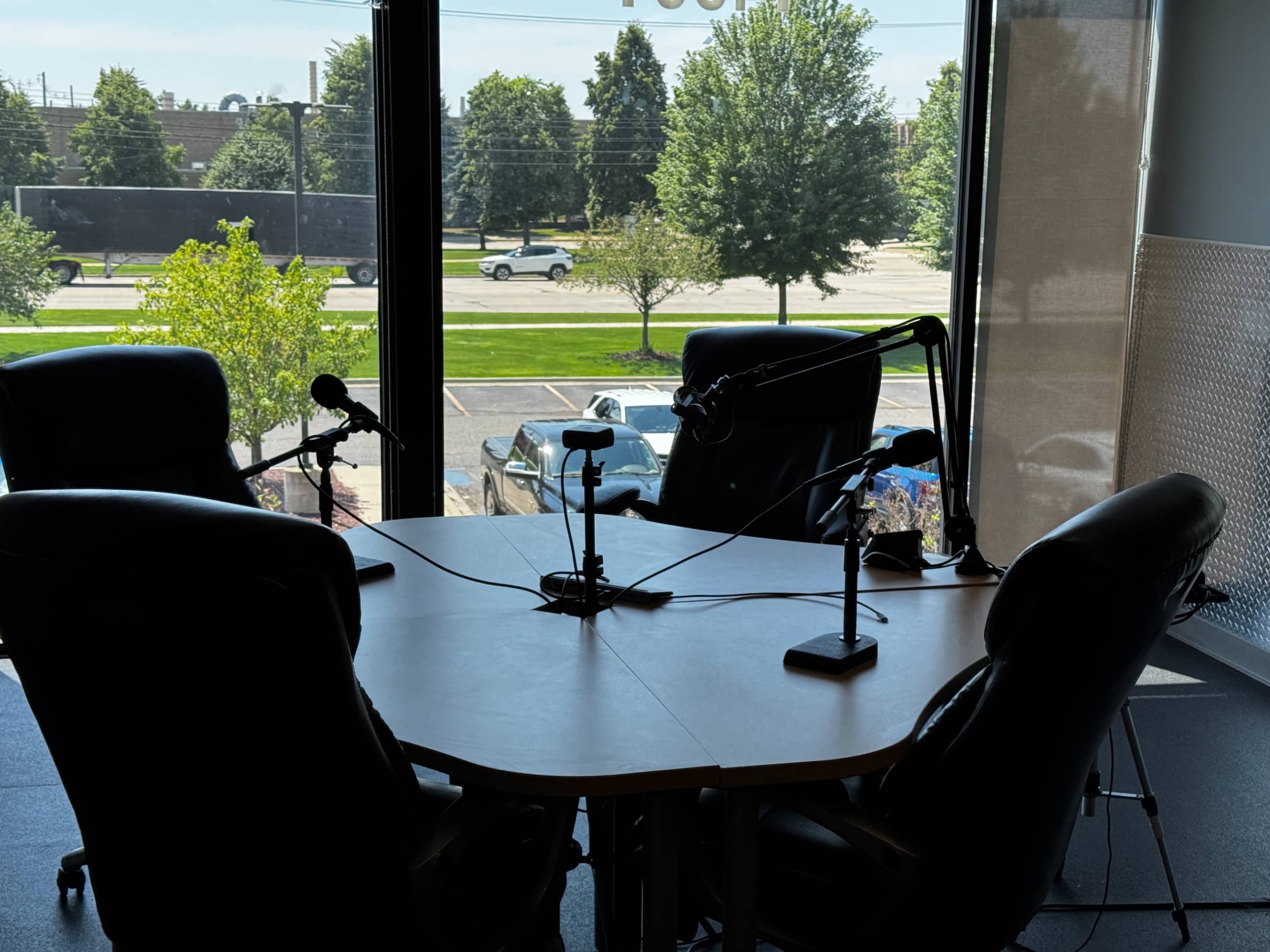The image shows a podcast recording setup with four black chairs and microphones positioned around a round table, facing large windows with a view of trees and a parking lot outside.