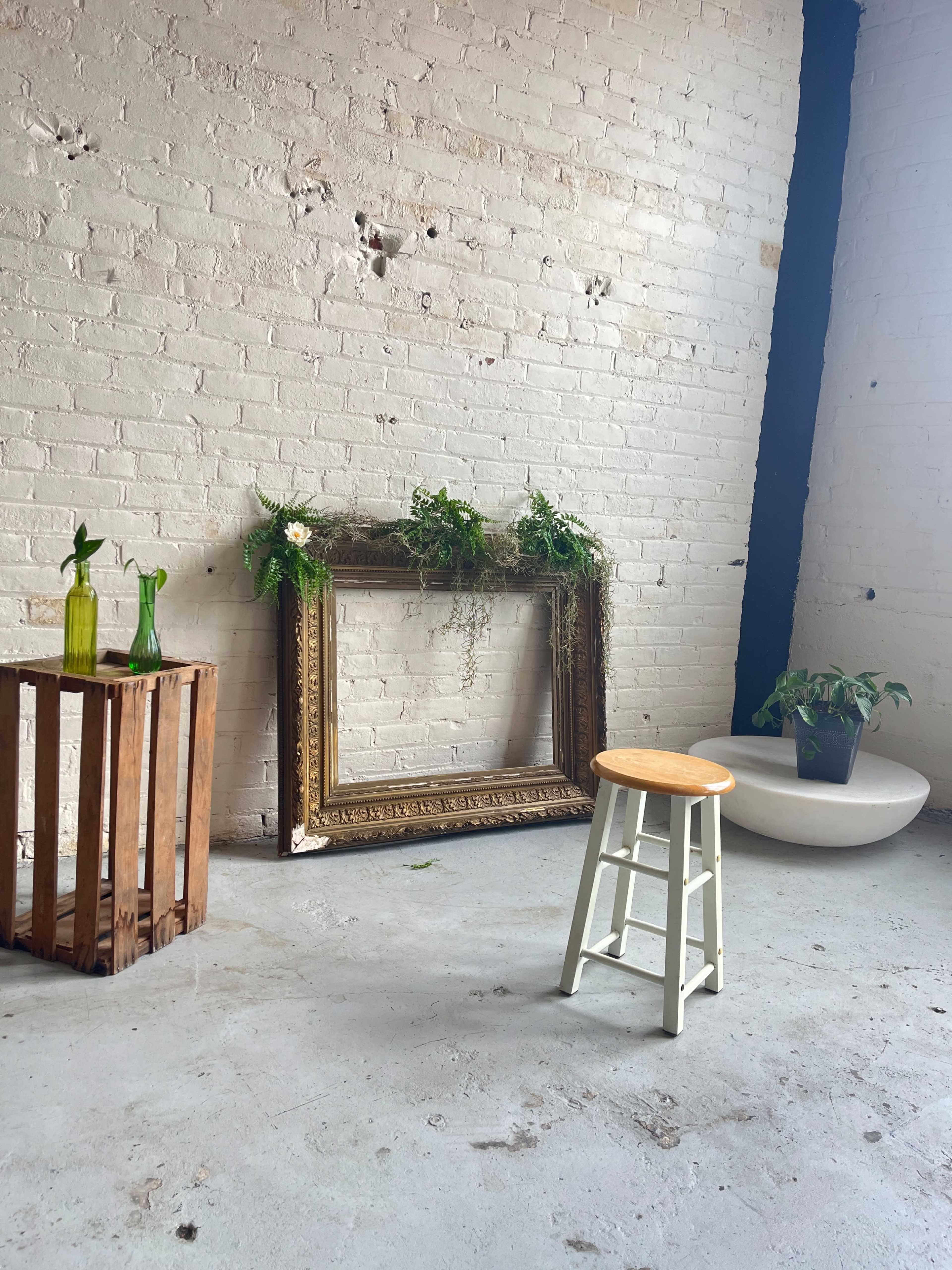 The room features a wooden stool and a vintage, ornate frame adorned with greenery, positioned against a white brick wall alongside a wooden crate and a potted plant.