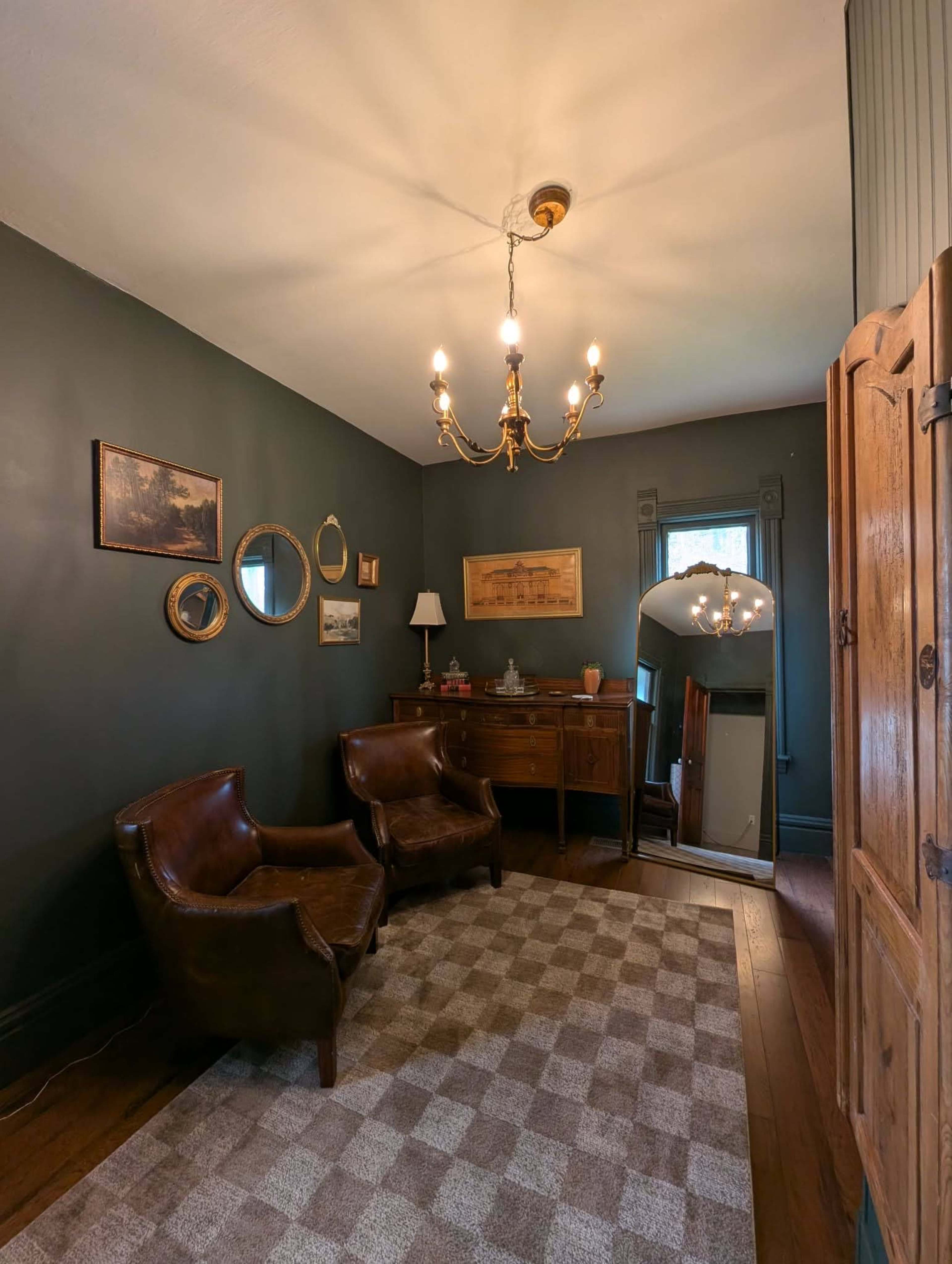 The image depicts a dimly lit, rustic room featuring two brown leather chairs, a wooden sideboard, a mirror, and various framed artworks on the walls.