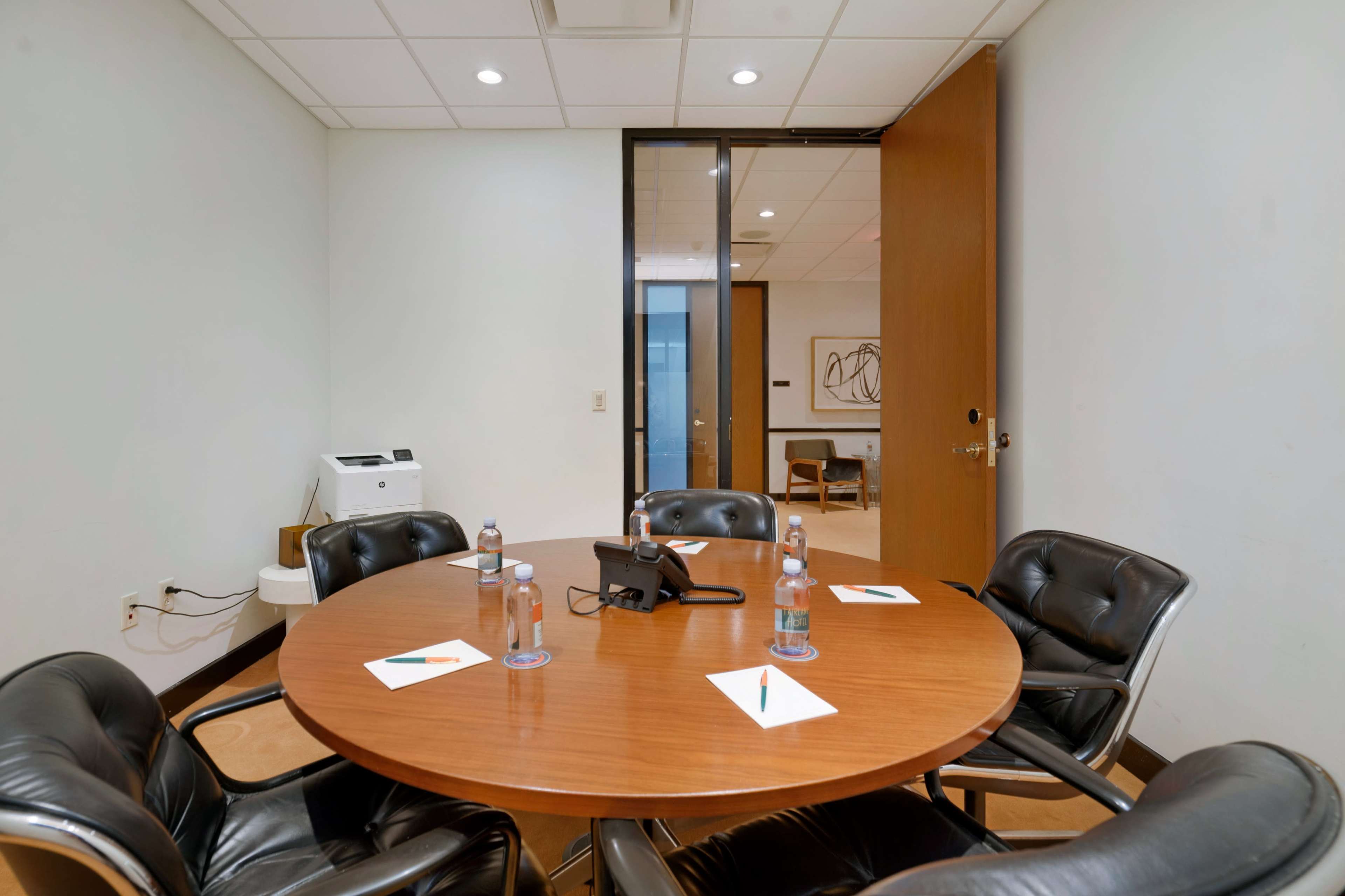 A round wooden table with black chairs and a phone is set up in a conference room, featuring water bottles and notepads at each seat.