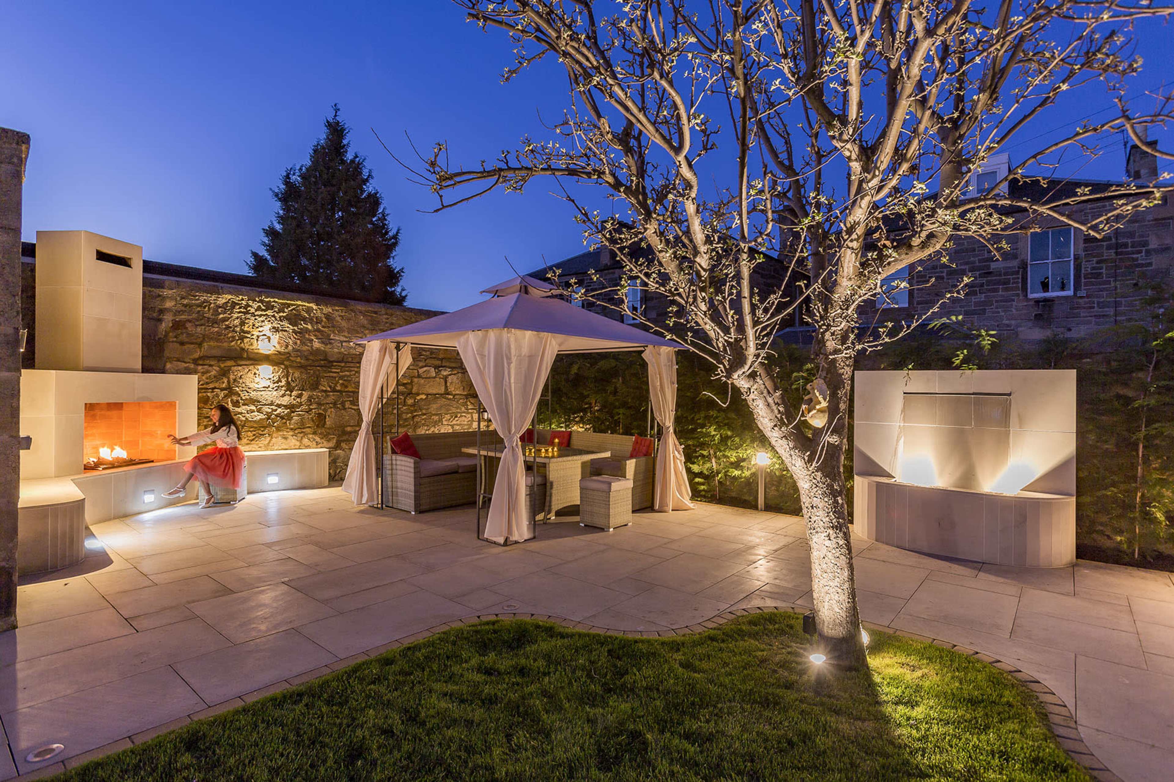 A landscaped outdoor area features a seating arrangement under a canopy, a stone fire pit, and a flowering tree.