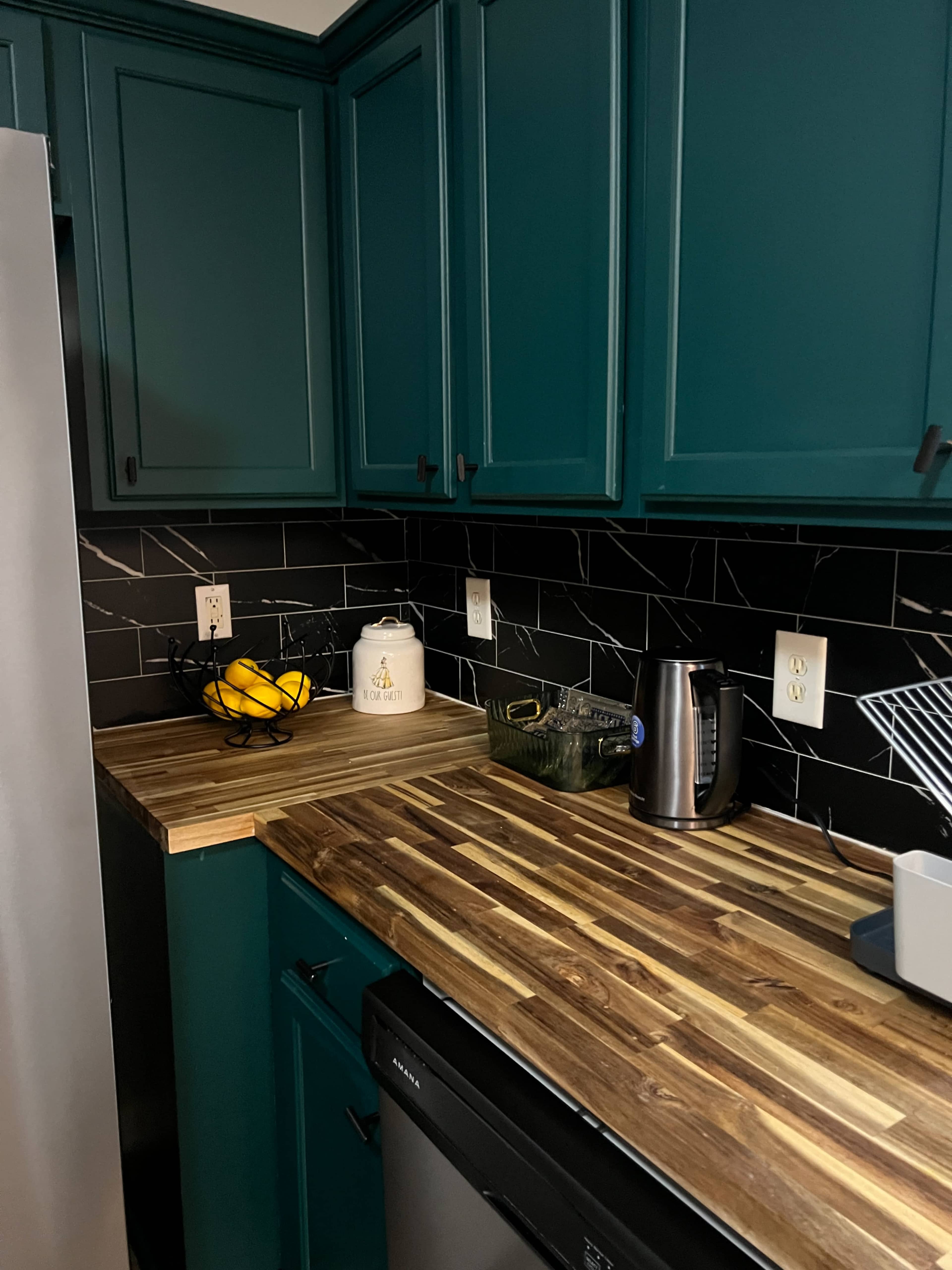 The image shows a kitchen with teal cabinets, a wooden countertop, and appliances including a kettle and a dish rack, against a black tiled backsplash.