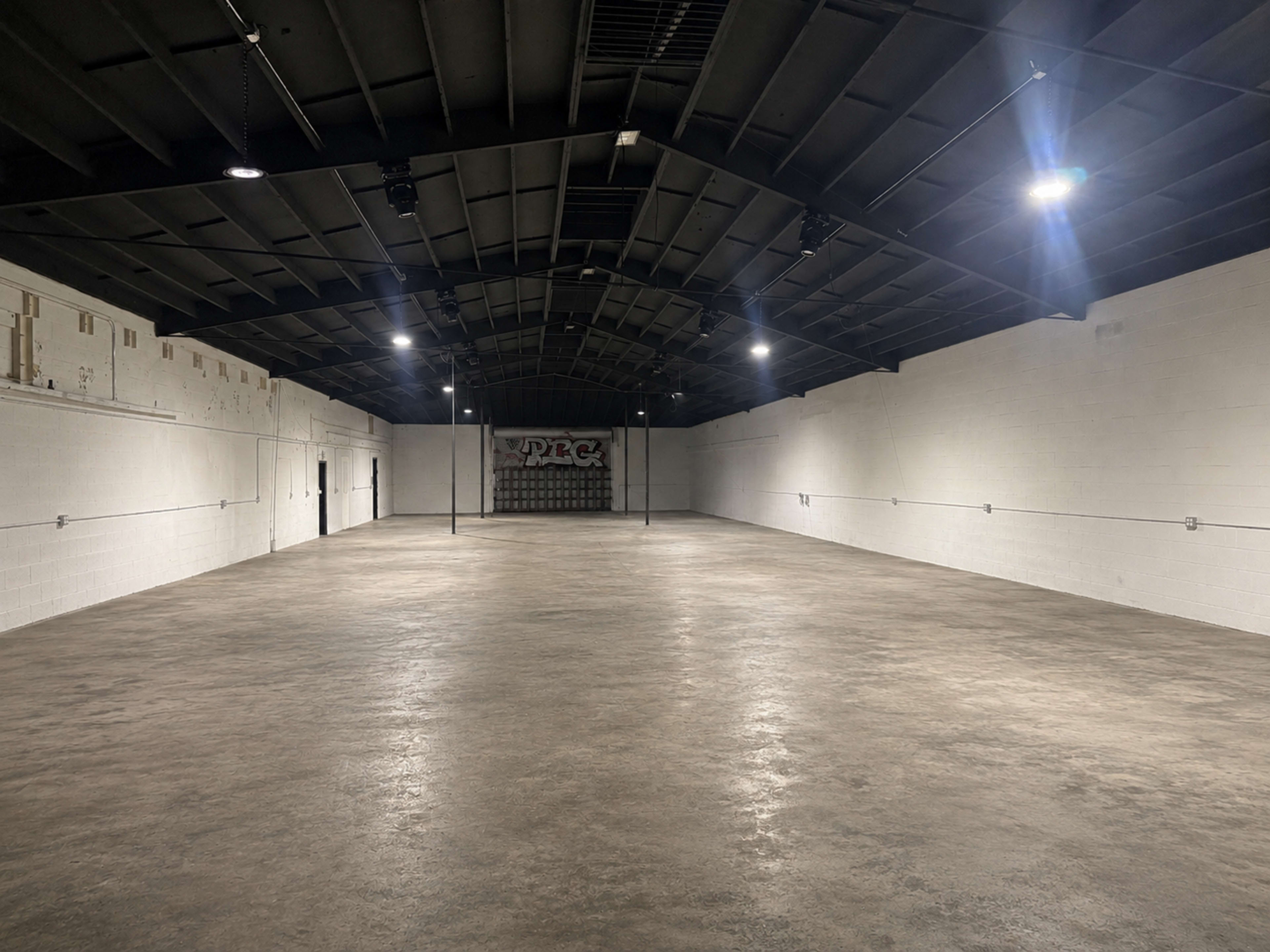 The image shows a large, empty warehouse space with a concrete floor and high, black ceiling, illuminated by overhead lights.