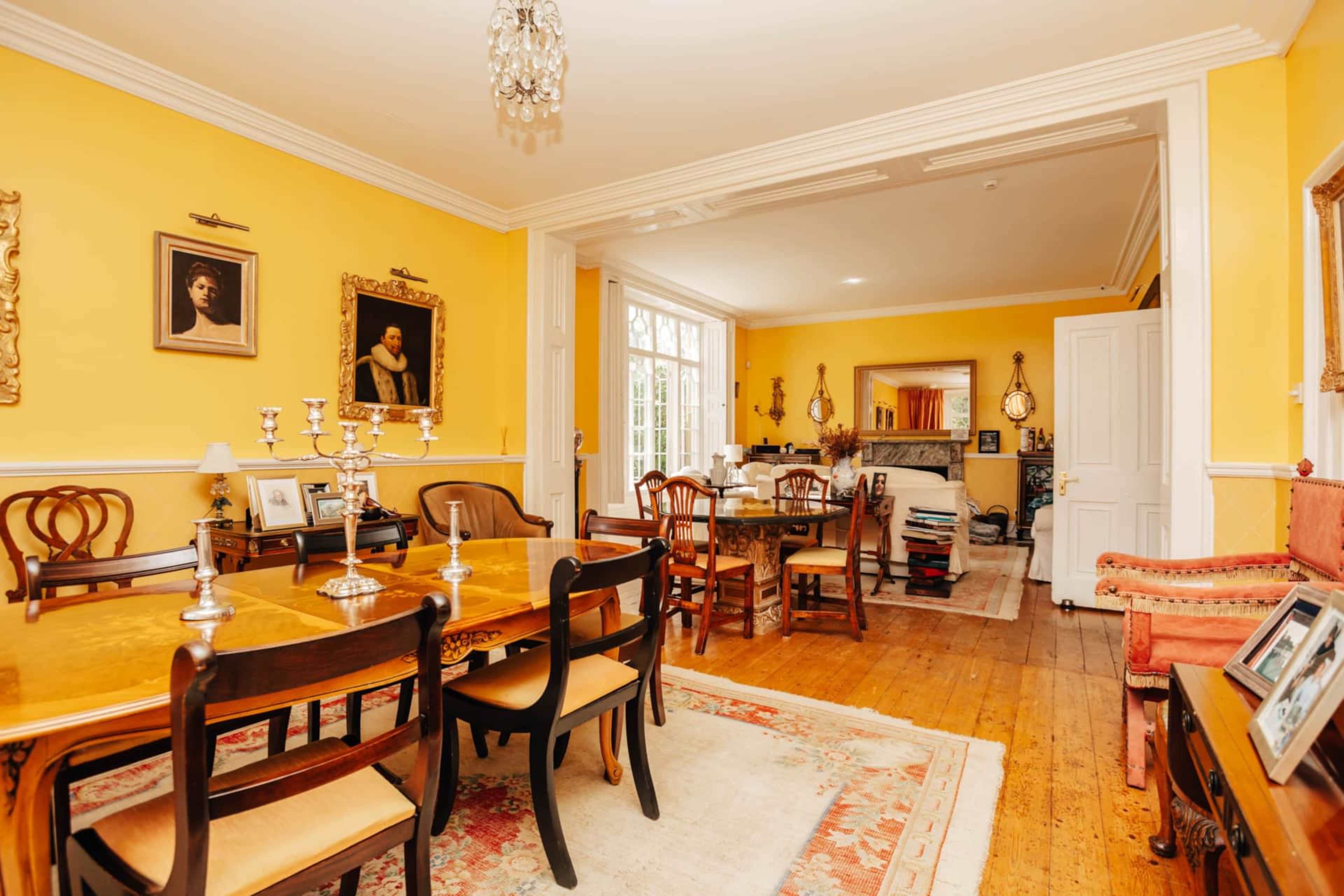 A brightly lit dining room featuring a wooden dining table surrounded by chairs, with yellow walls adorned with framed portraits and a chandelier overhead.