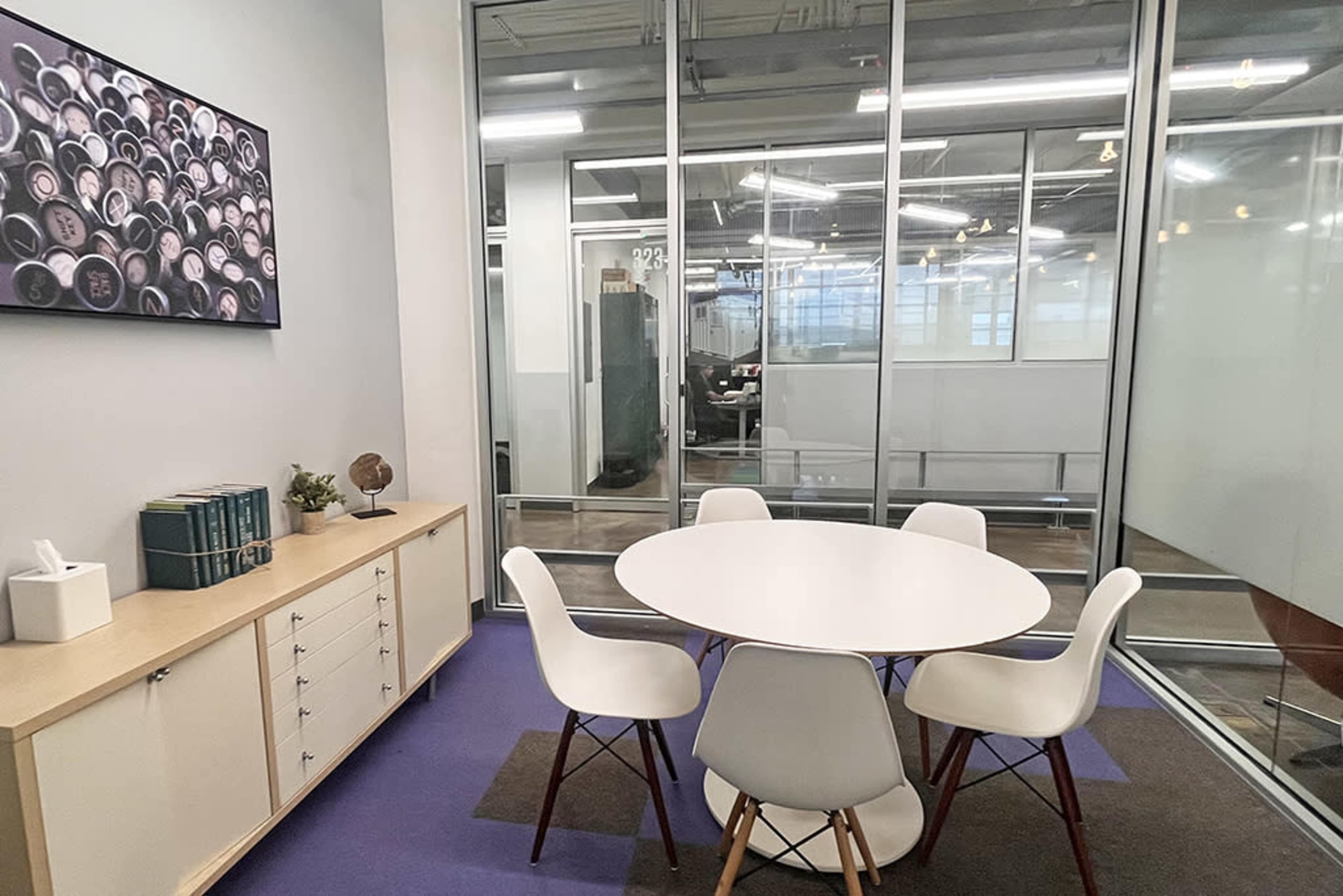 The image shows a small meeting room with a round table surrounded by four white chairs, a cabinet with books and a globe, and glass walls providing a view of an office space beyond.