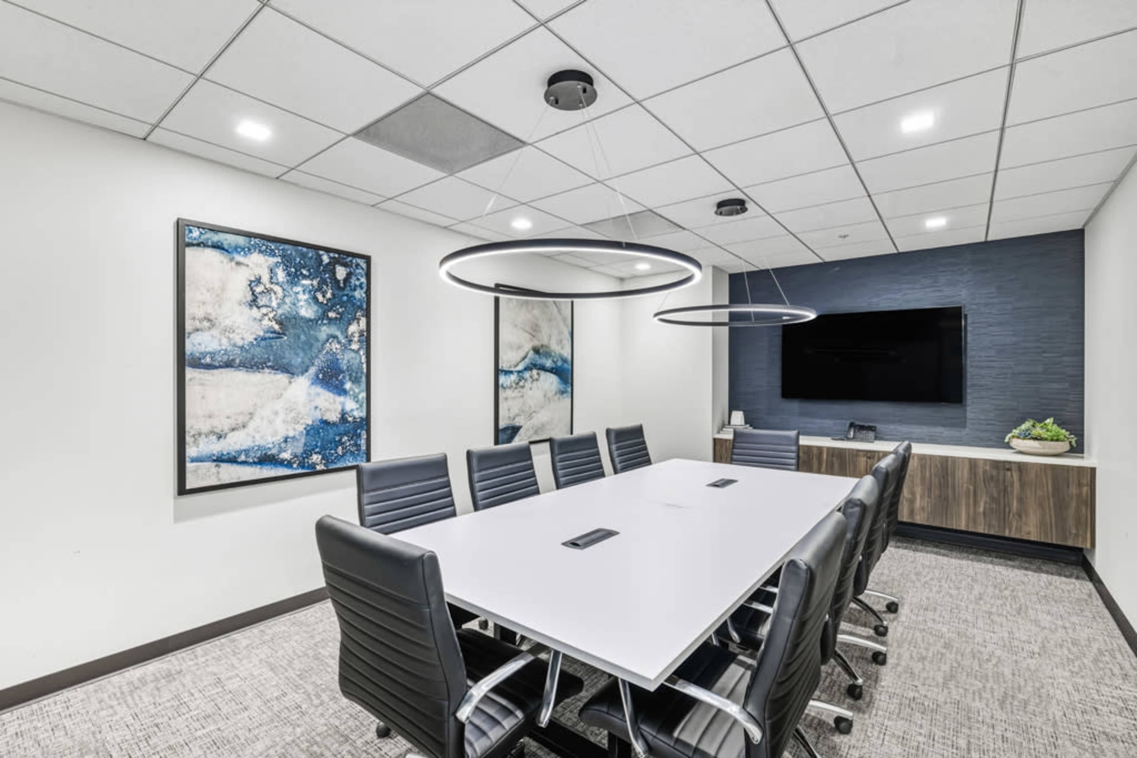The image shows a modern conference room with a long white table, black leather chairs, two abstract paintings on the walls, and a television mounted above a wooden cabinet.