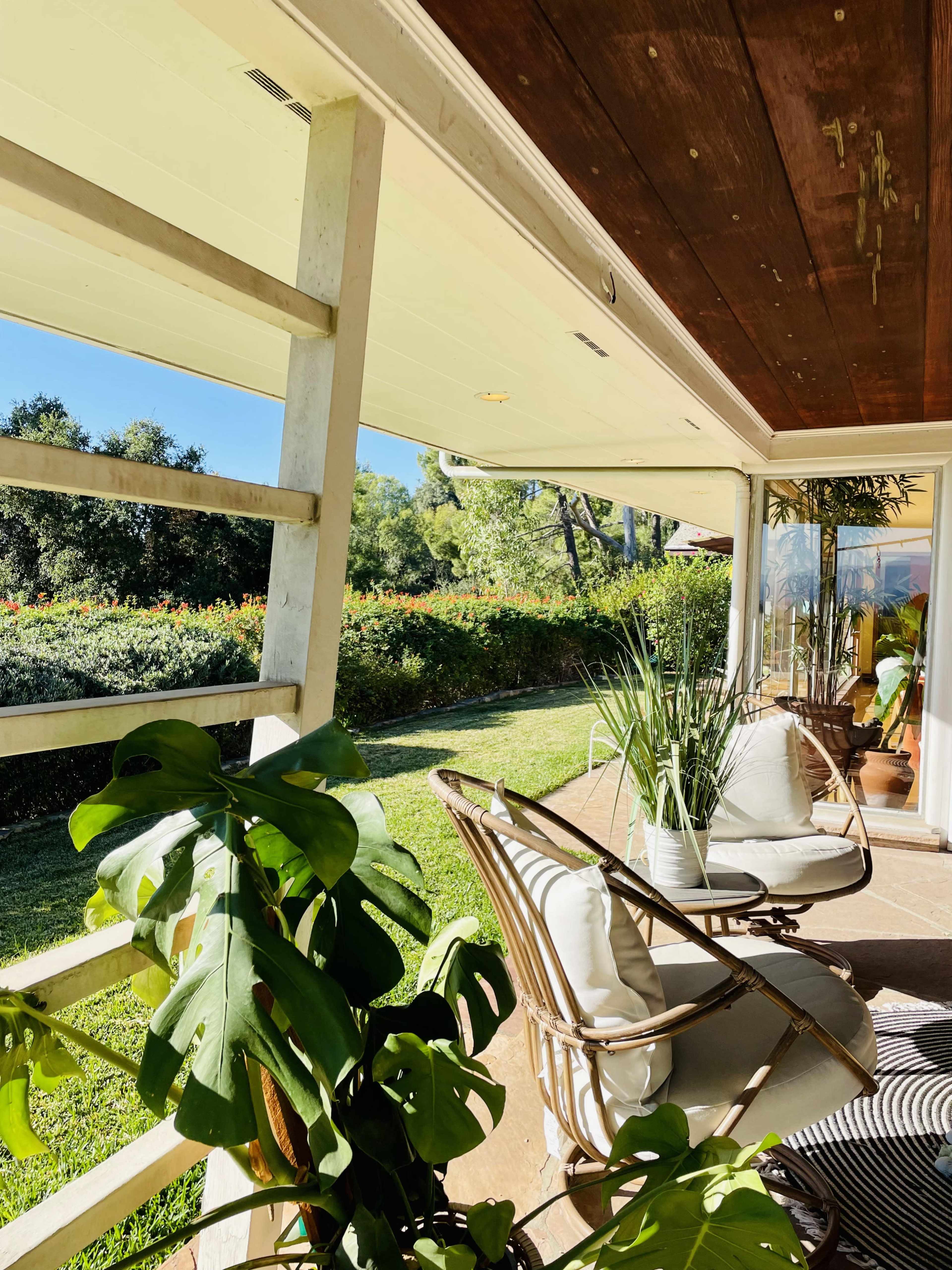 The image shows a porch with two white chairs, surrounded by lush greenery and a clear blue sky in the background.