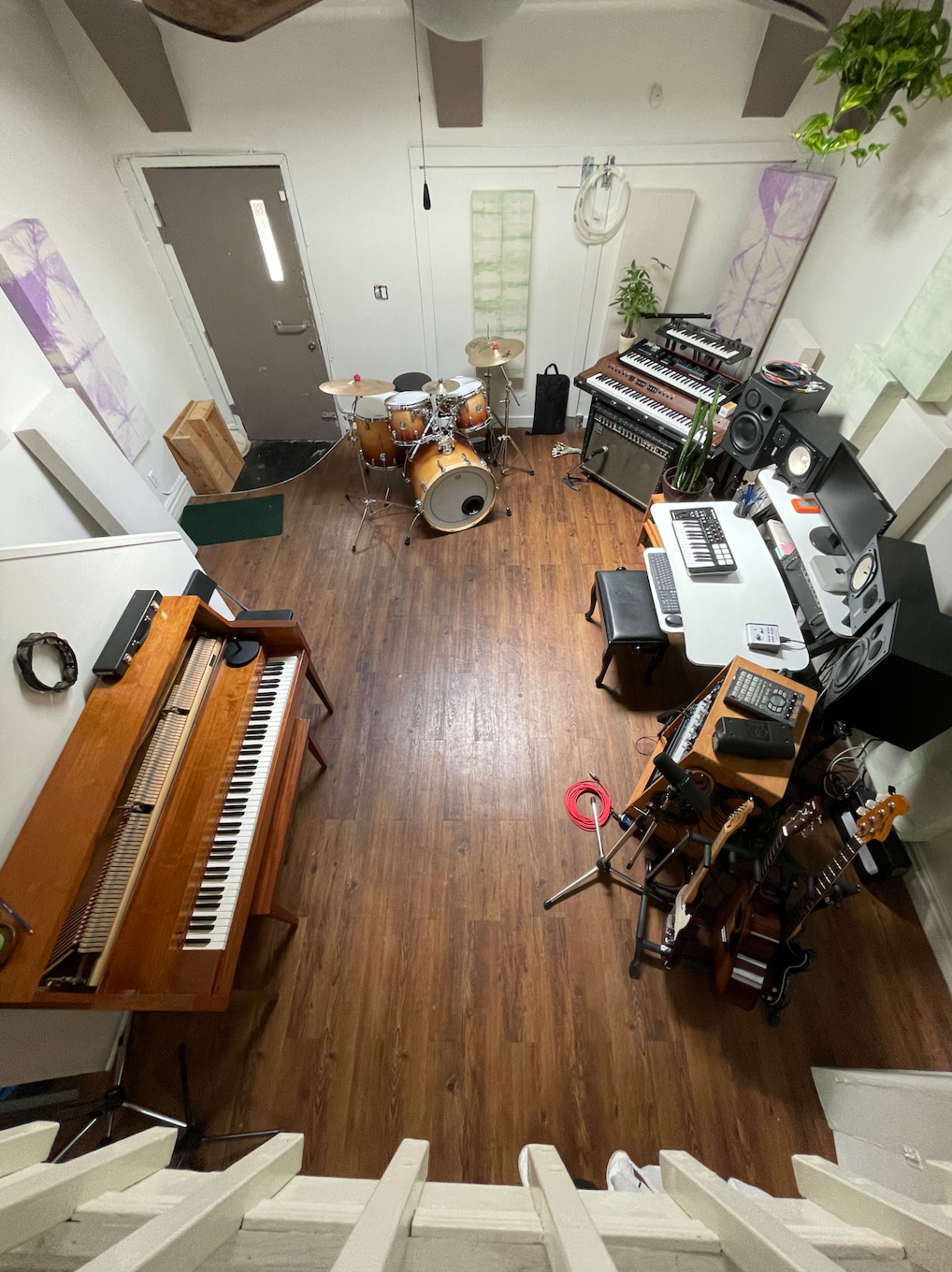A music studio featuring a drum set, a keyboard, synthesizers, and various guitars arranged in a wooden-floored room.