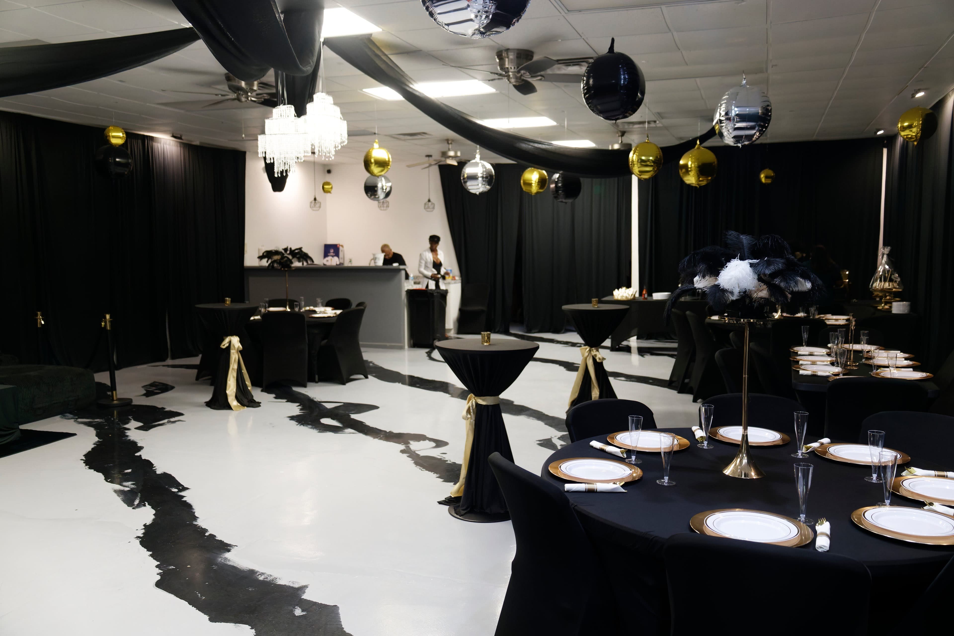 The image shows a decorated event space with black tables, gold accents, and hanging decorations, set up for a formal gathering.