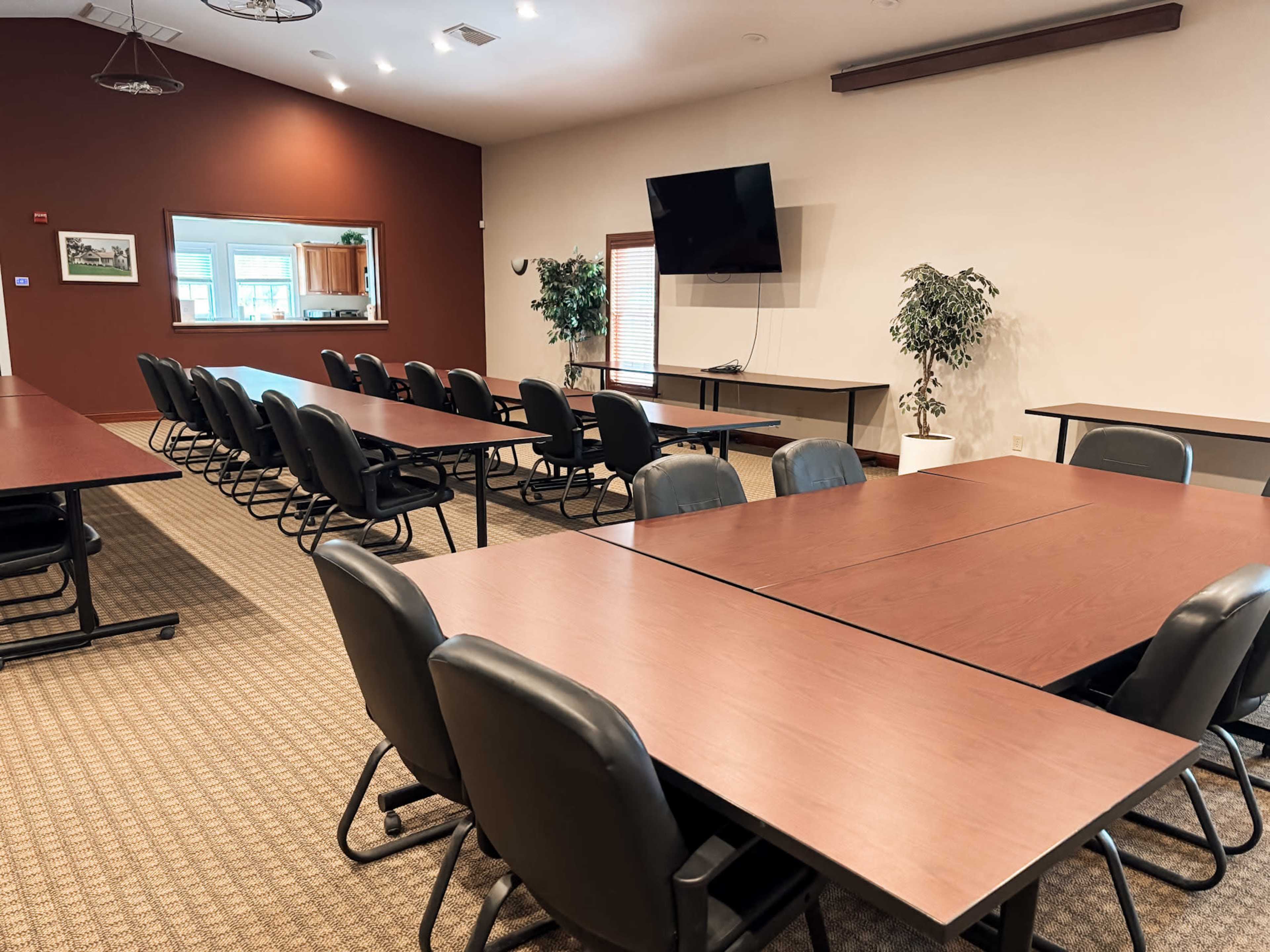 The room features several rows of rectangular tables with black chairs, a large television screen on the wall, and a small kitchen area visible in the background.