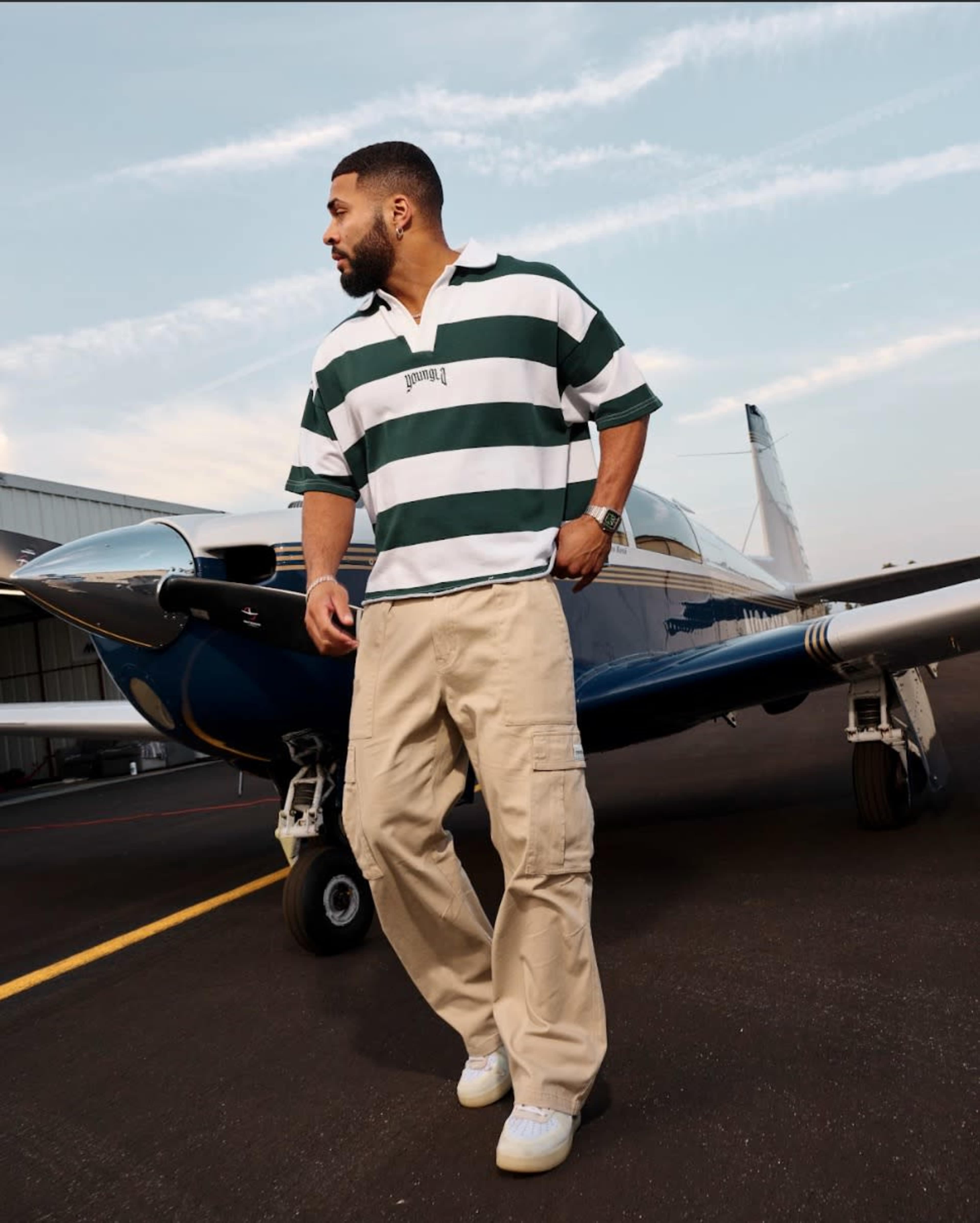 A man in a striped polo shirt and beige cargo pants stands next to a small aircraft on an airstrip.