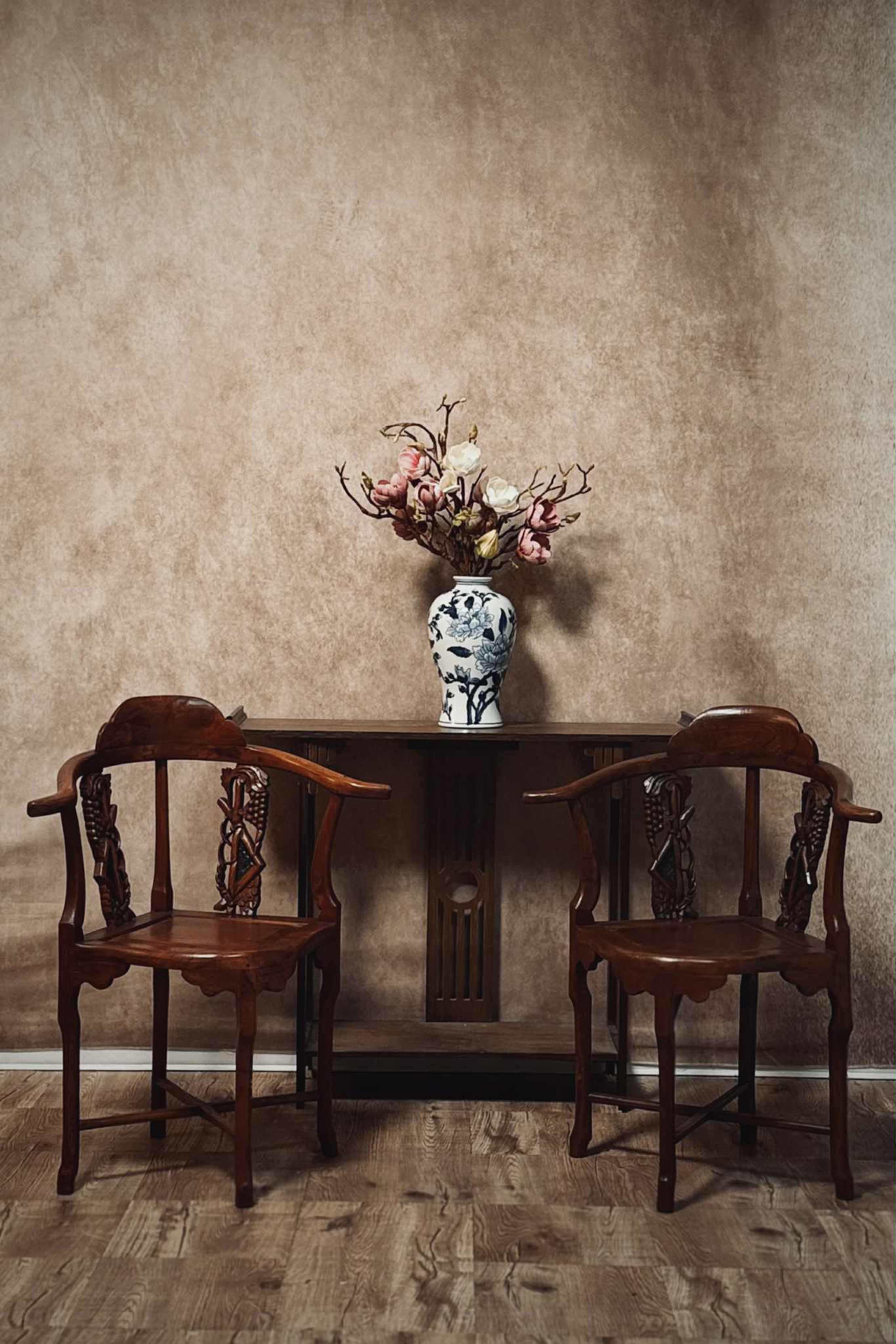 A wooden table with two matching chairs is positioned against a textured wall, topped with a vase containing flowers.