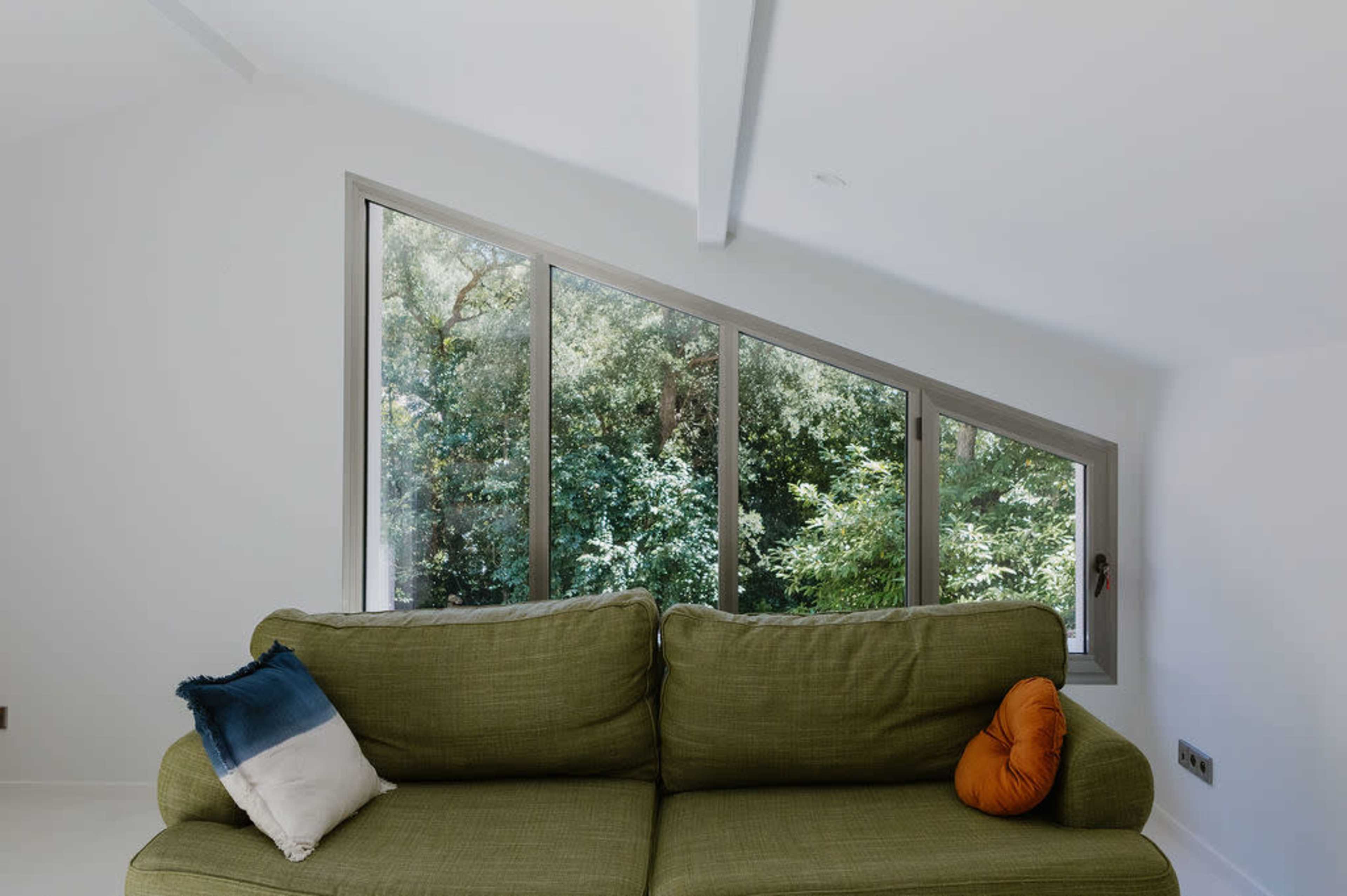 A green couch with orange and blue cushions is positioned beneath large angular windows that frame a view of lush greenery outside.