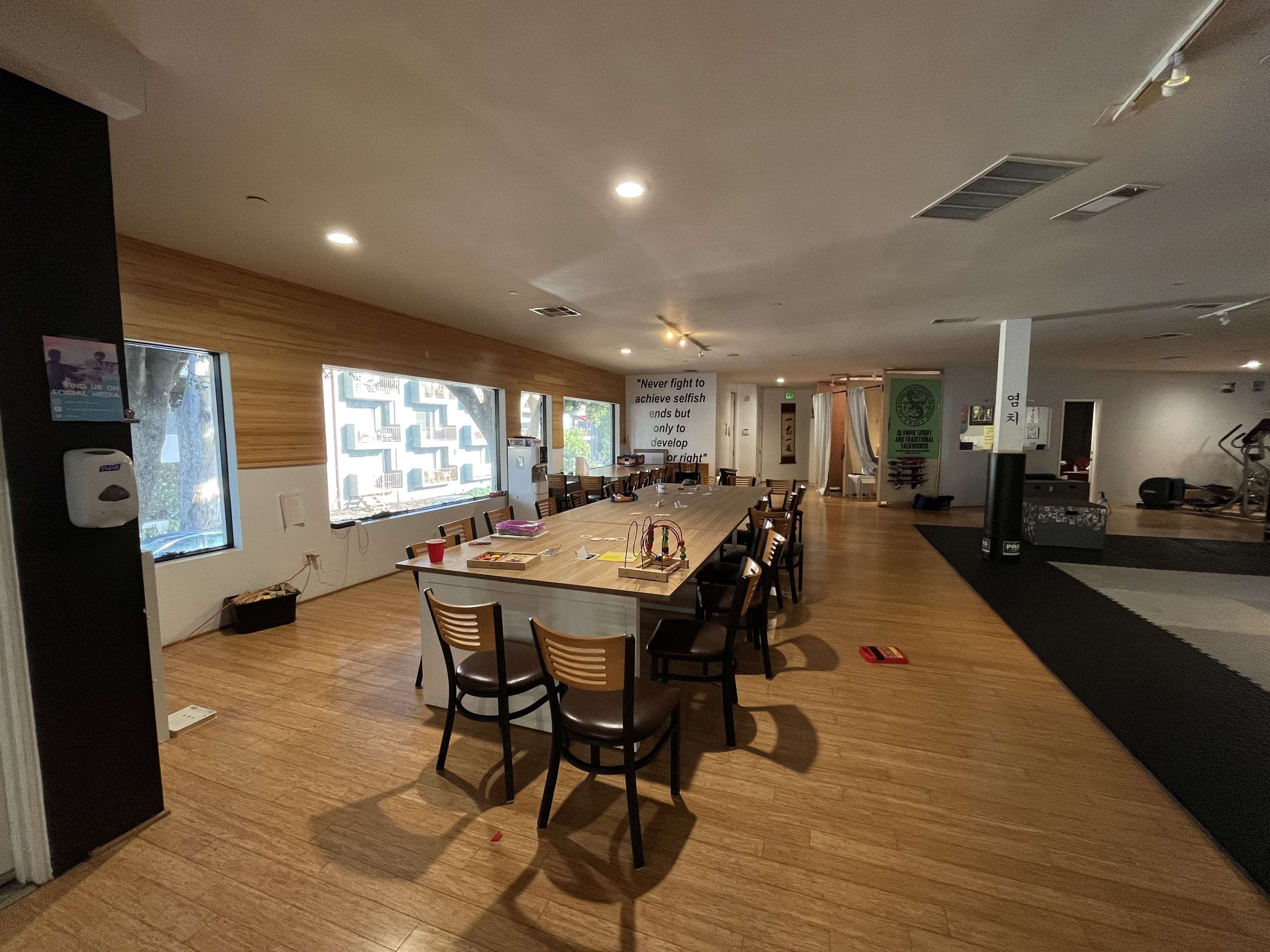 A spacious room furnished with a long table surrounded by chairs, with large windows on one side and exercise equipment visible in the background.