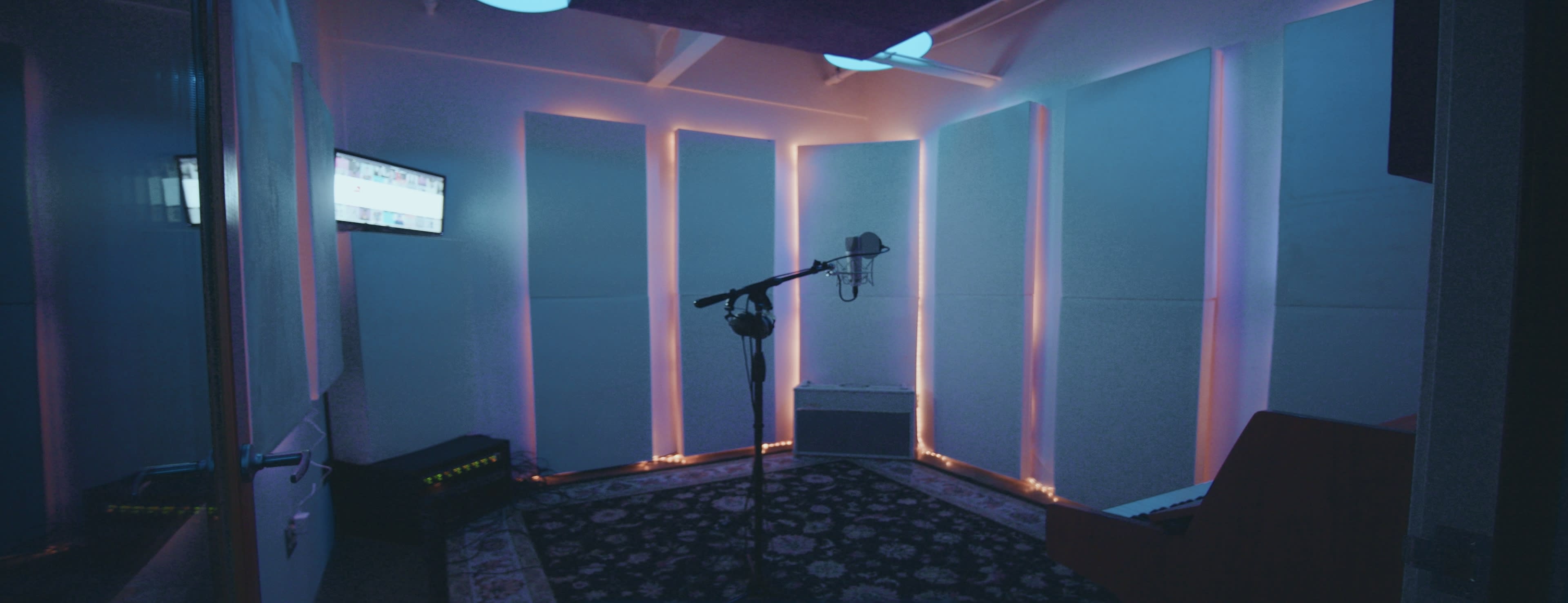 A sound recording studio features a microphone on a stand, blue and purple lighting along the walls, and sound-absorbing panels.