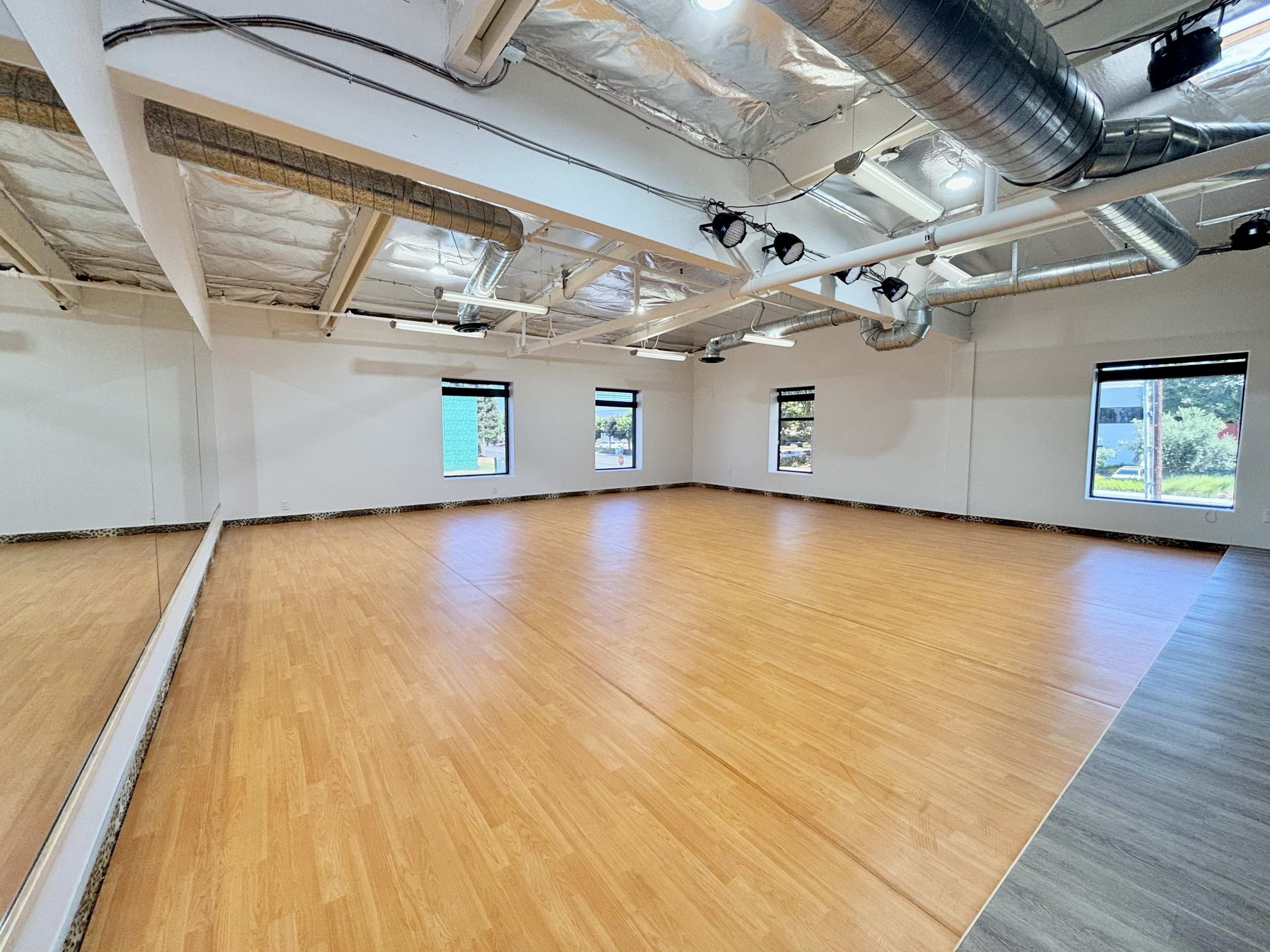 The image shows a spacious, empty studio with wooden flooring, bright white walls, large windows, and a mirrored wall reflecting the room's interior.