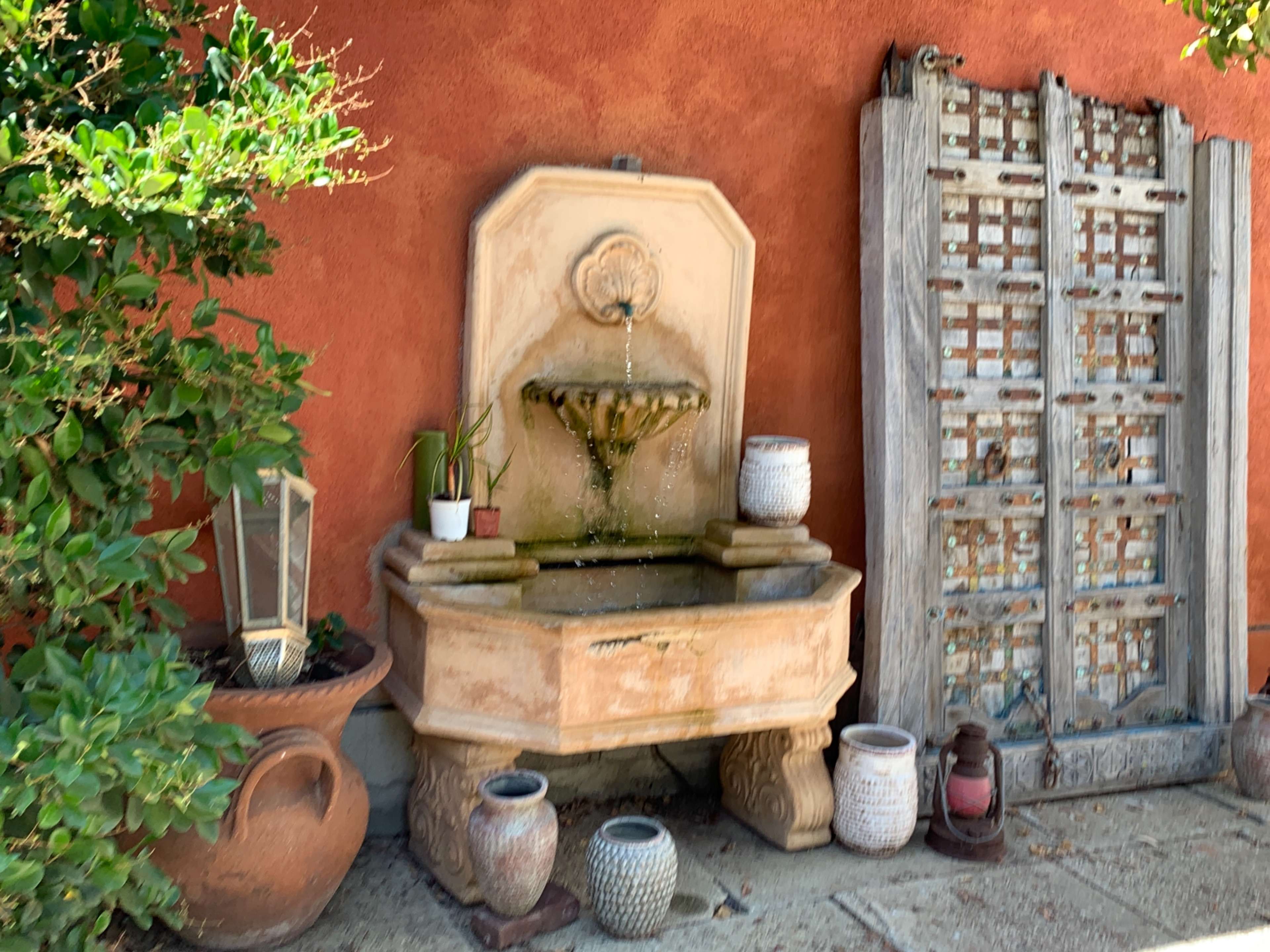 A decorative stone fountain with water flowing into a basin is positioned against an orange wall, accompanied by potted plants and various containers nearby.