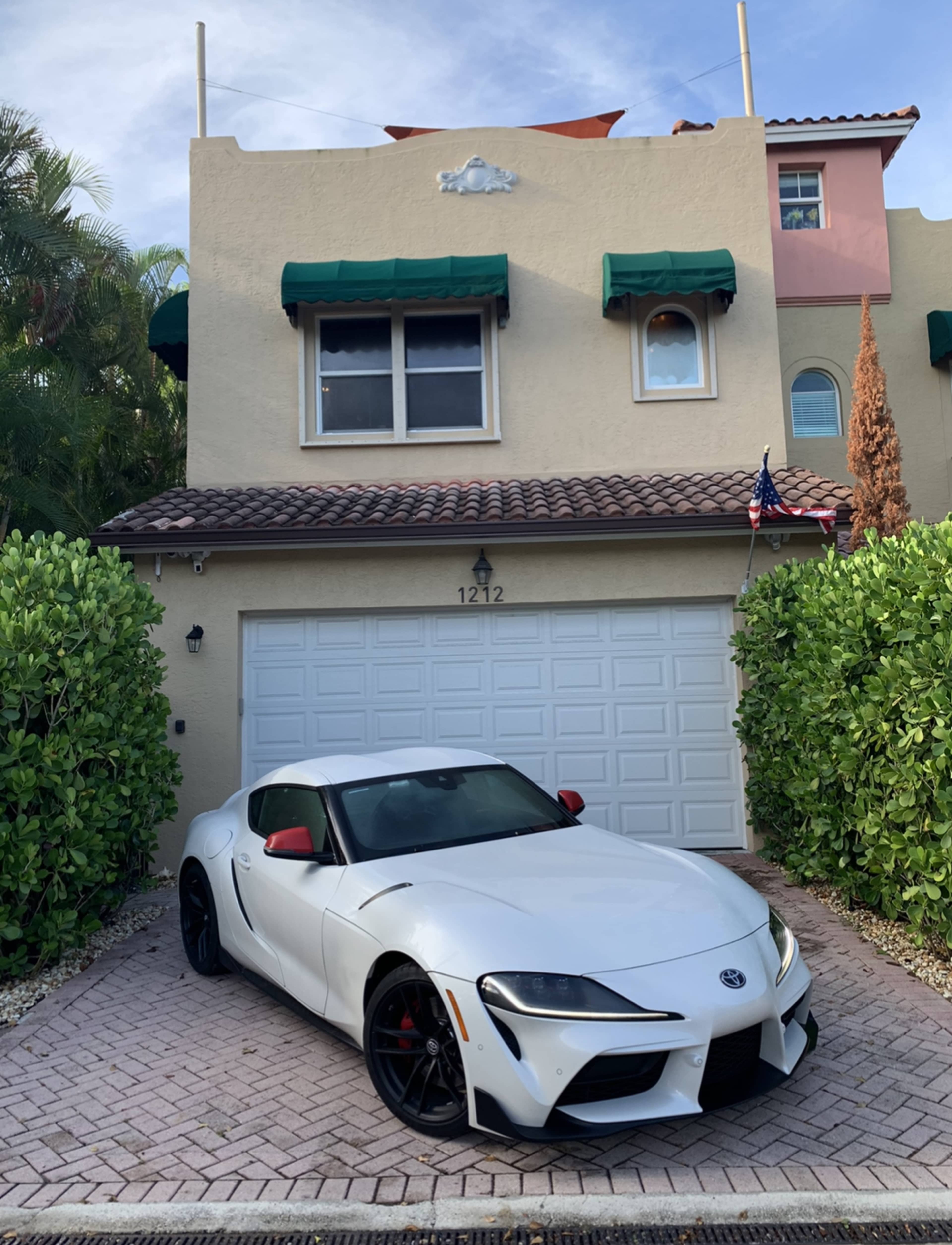 A white Toyota Supra is parked in front of a two-story, beige house with green awnings and a garage door.