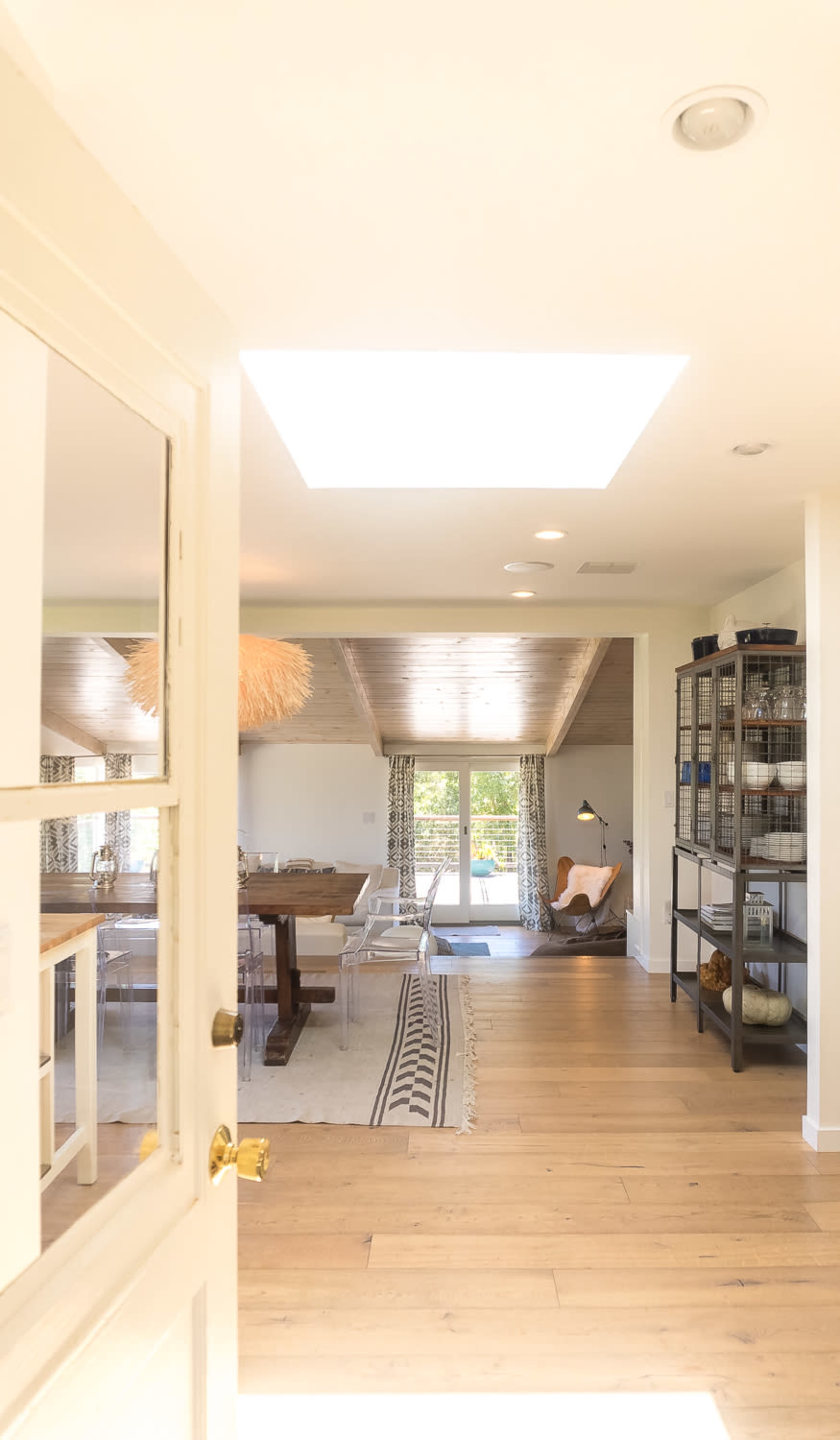 The image shows a well-lit interior space with wooden flooring, a dining table, and a view of another room featuring a large window and decorative shelves.