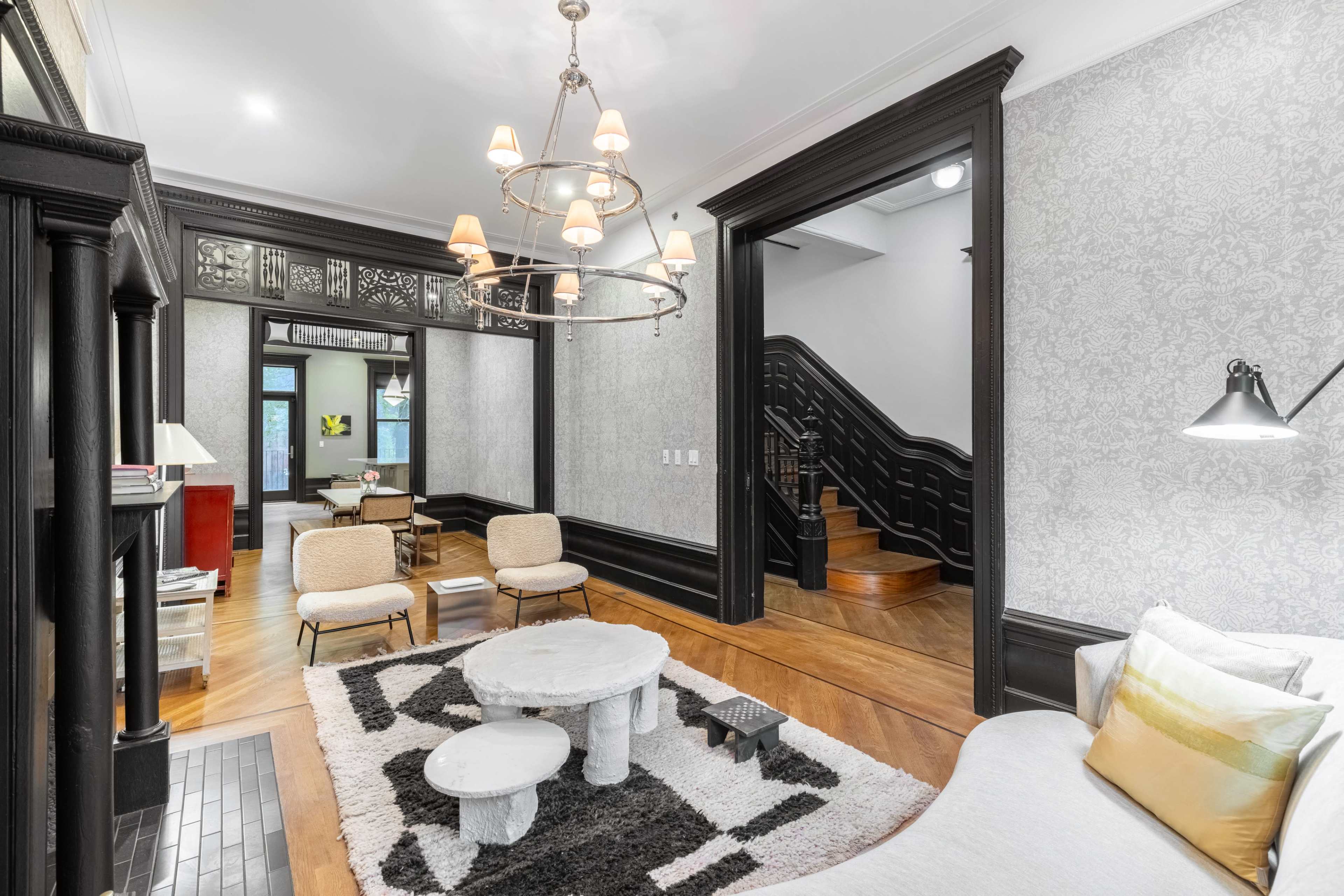 A stylish living room featuring a light-colored sofa, two chairs, a round coffee table on a patterned rug, and a staircase in the background.