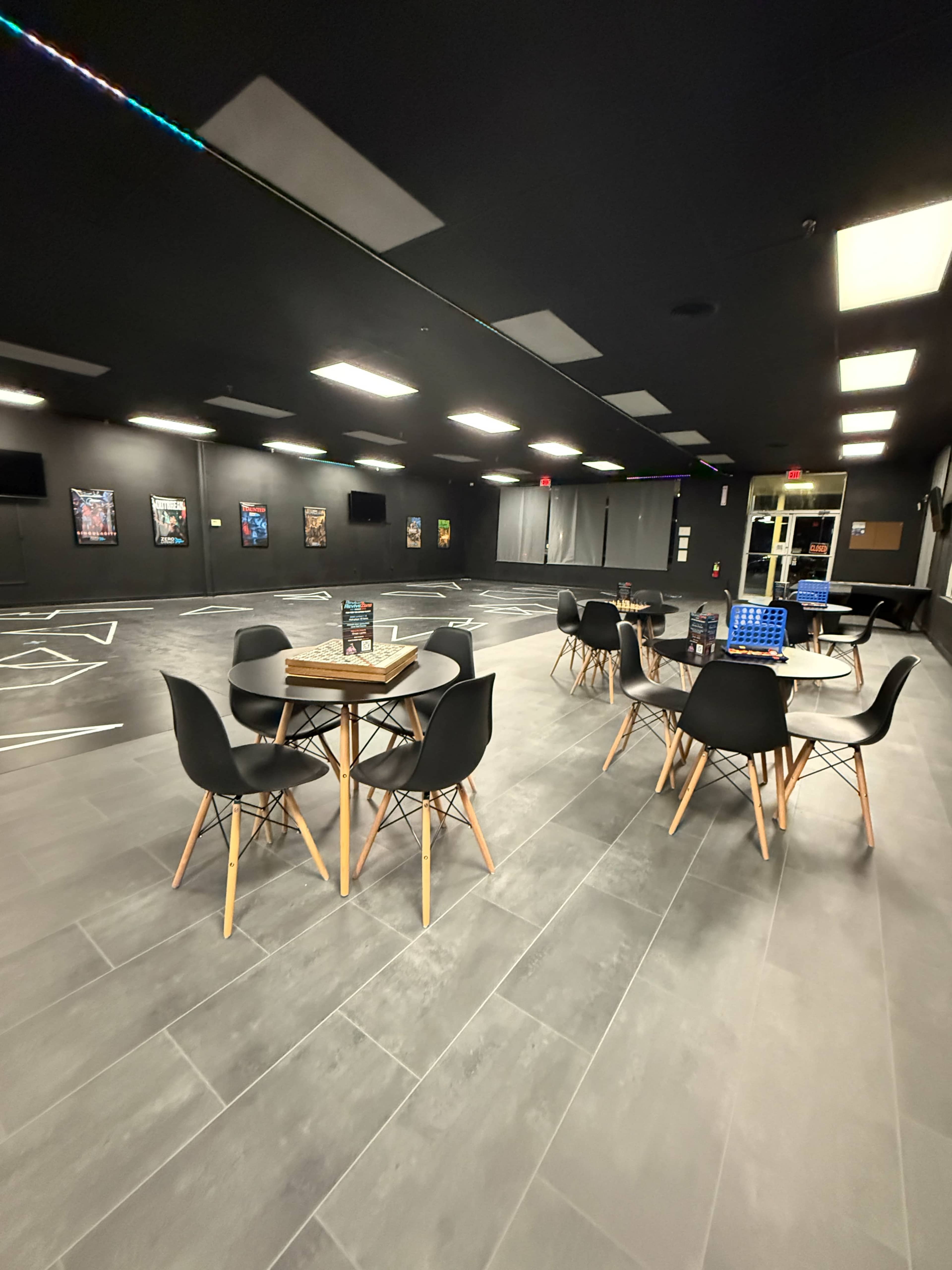 The image shows a spacious, dimly lit room with several tables and chairs arranged on a tiled floor, accompanied by wall posters and ceiling lights.