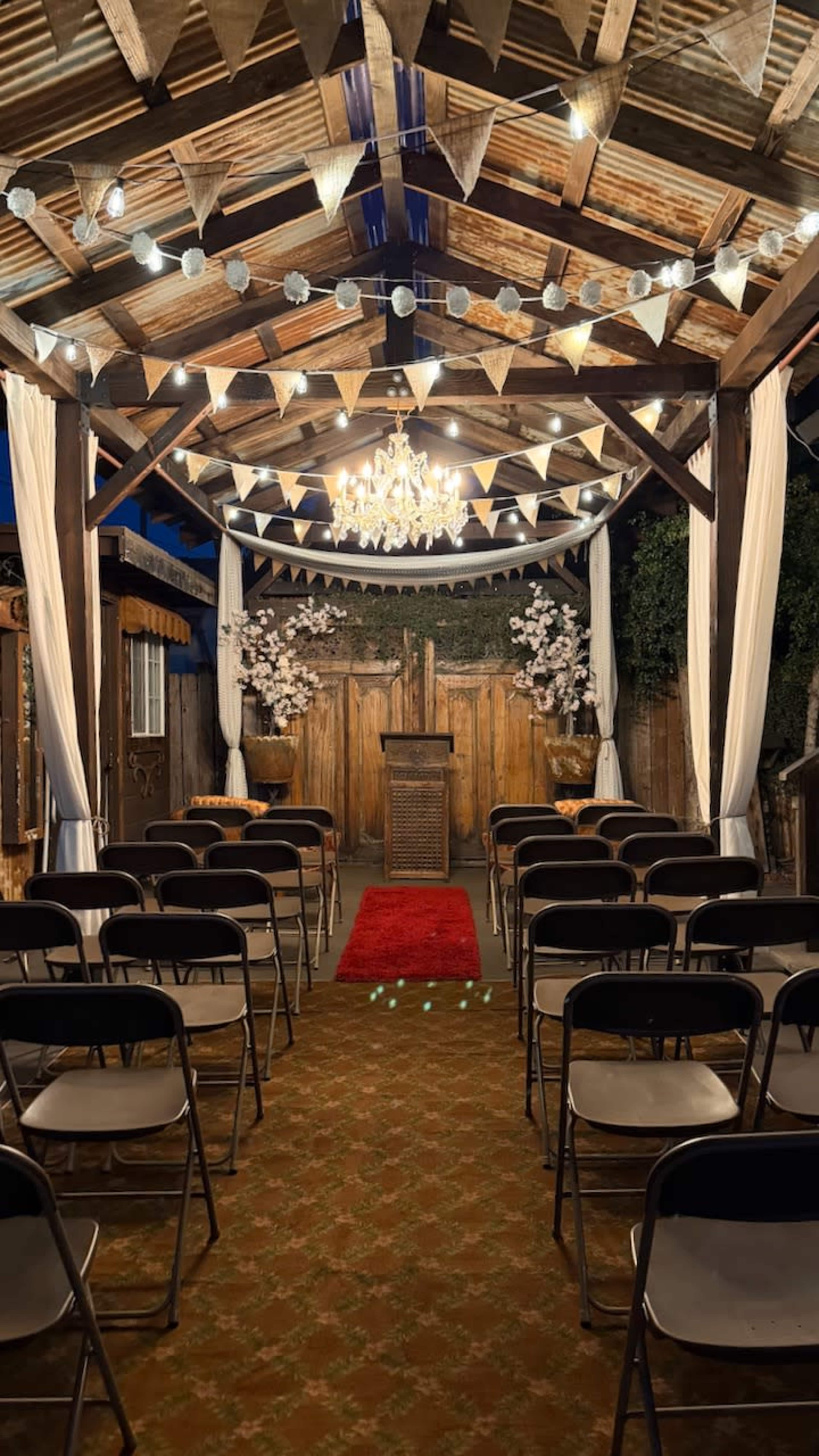 The image shows a rustic indoor ceremony space with rows of metal chairs facing a wooden altar decorated with floral arrangements and draped with bunting and lights.
