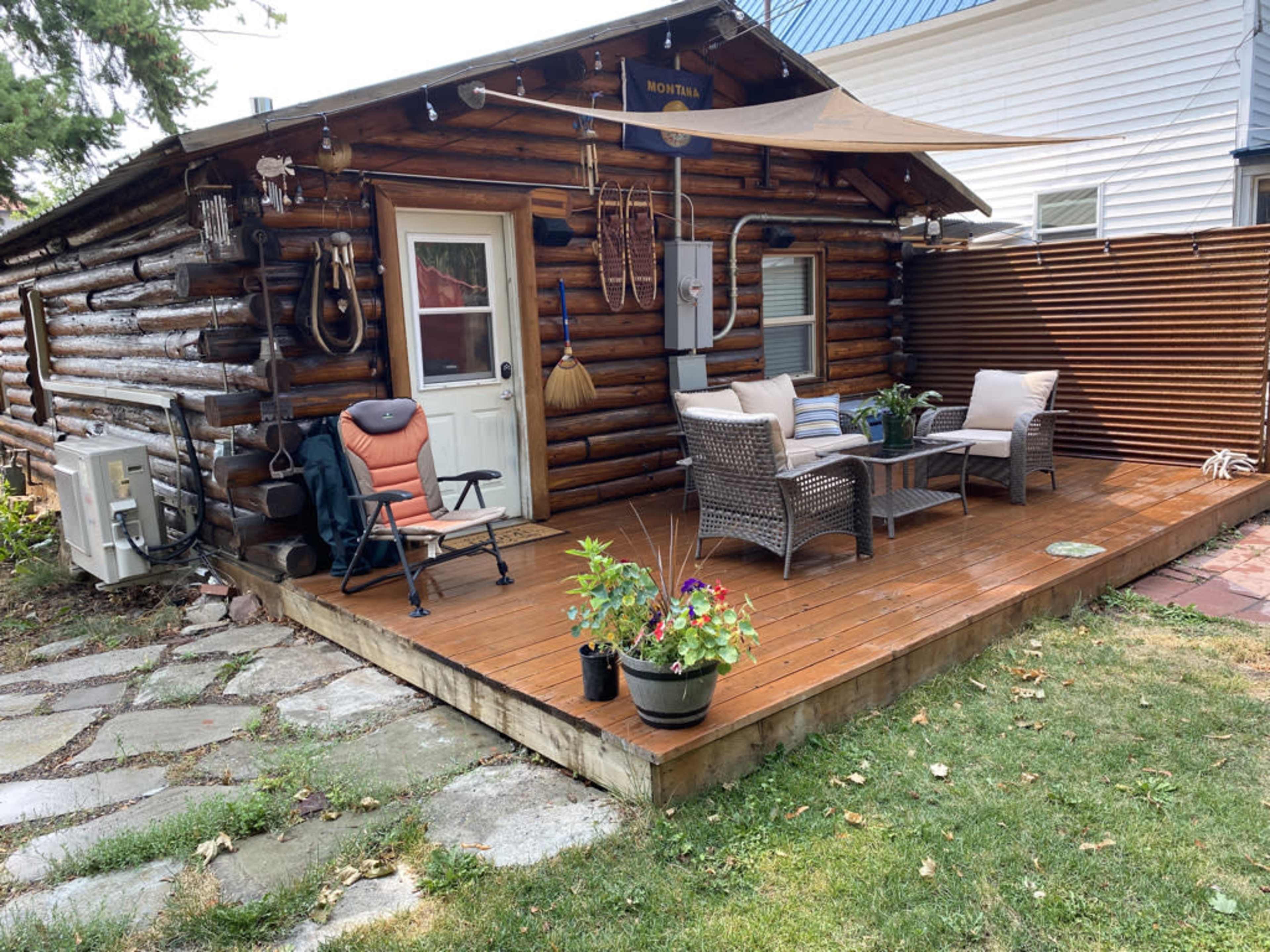 A wooden log cabin features a small deck with outdoor seating and potted plants surrounded by grass and stone pathways.