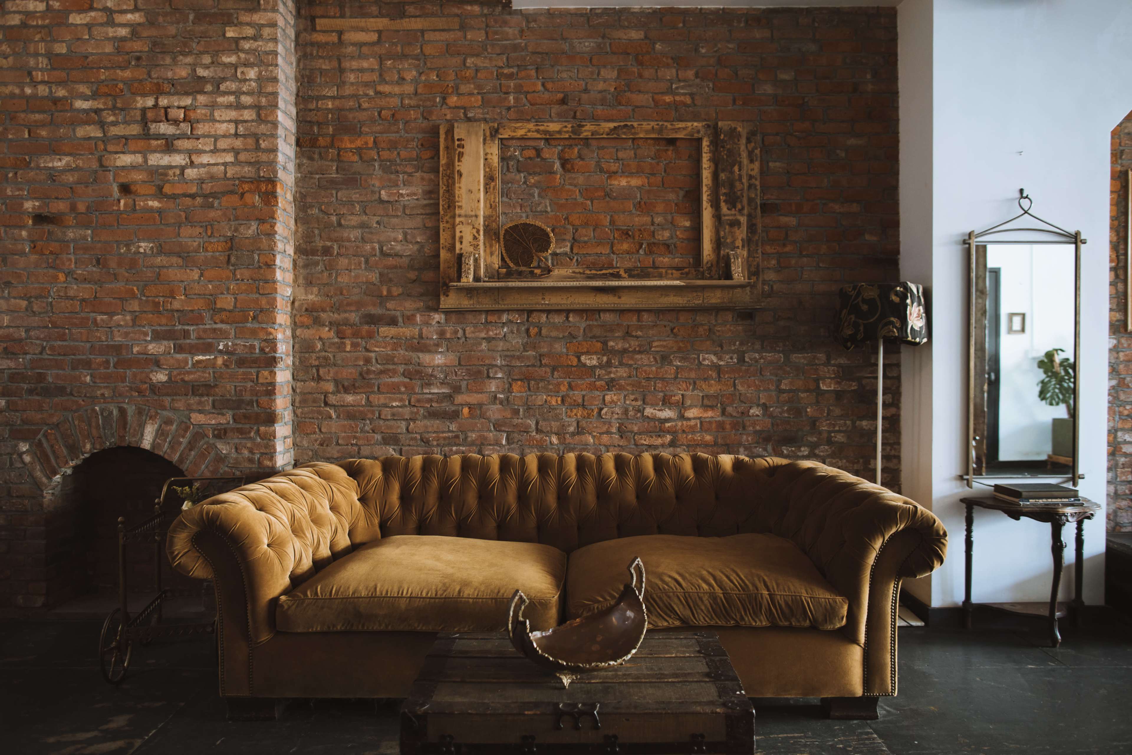 A brown tufted sofa is positioned against a brick wall, with a wooden frame and mirror accentuating the space.