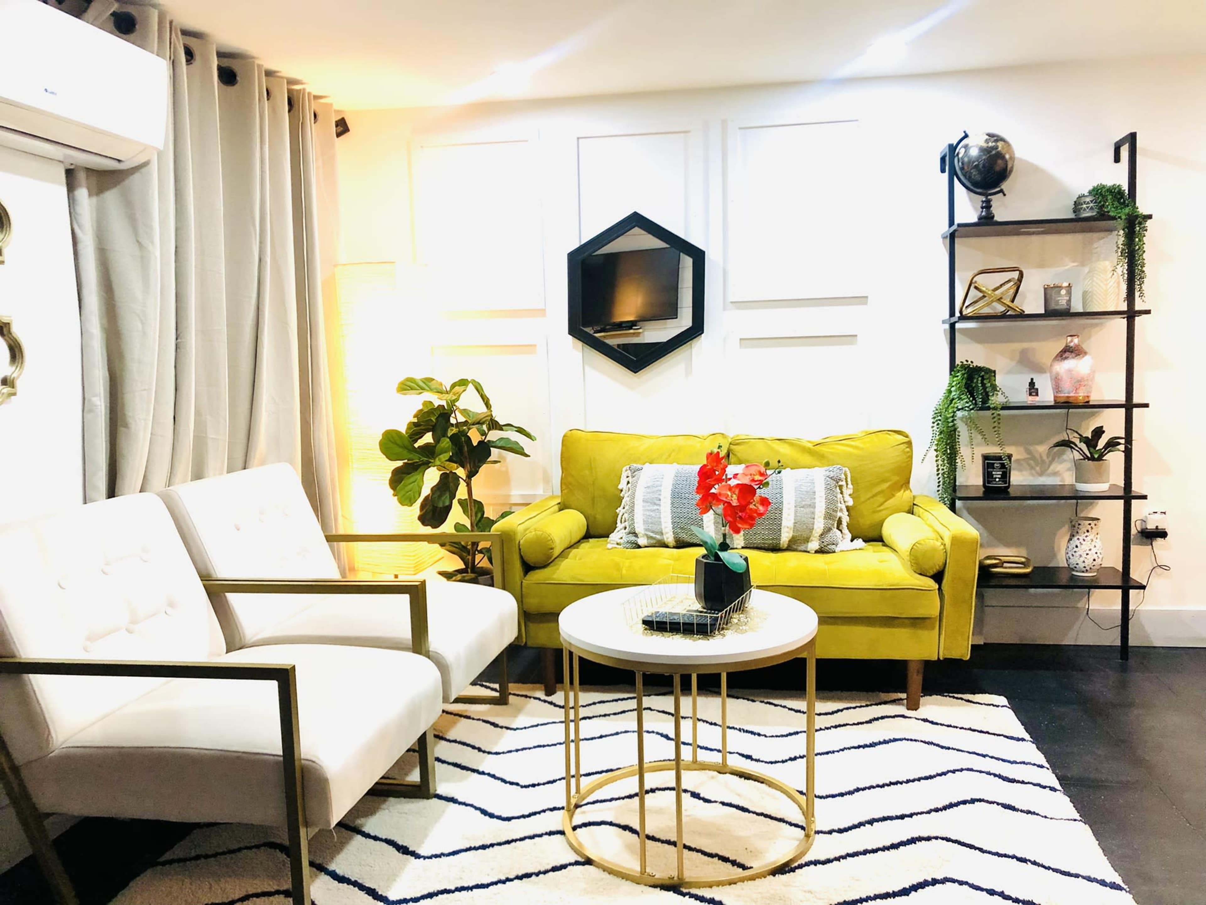 The image depicts a cozy living room featuring a green sofa, two white chairs, a round coffee table, and decorative shelves with plants and accessories.