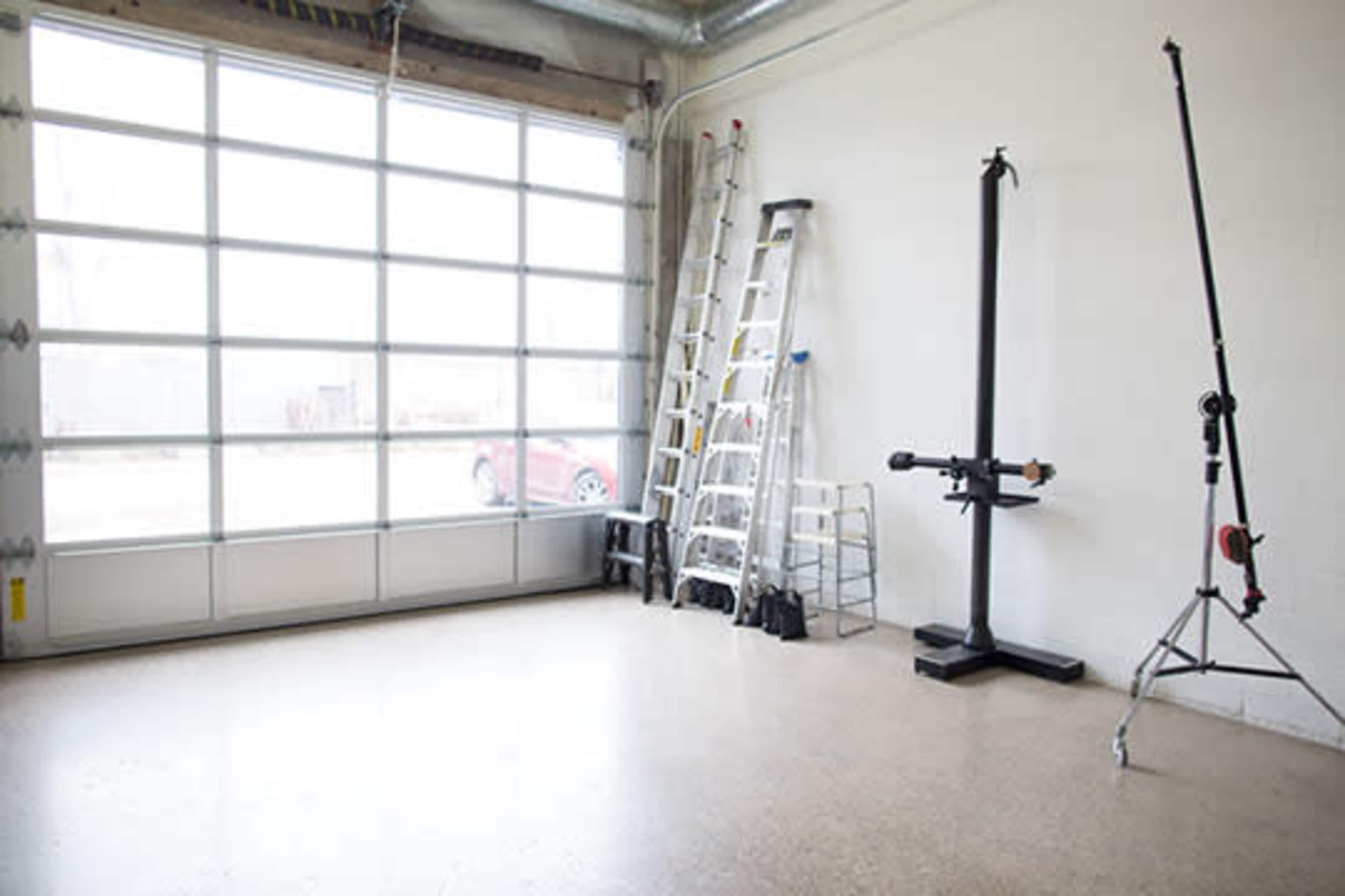 The image shows an empty garage or workshop space with ladders and a black equipment stand arranged along a white wall.