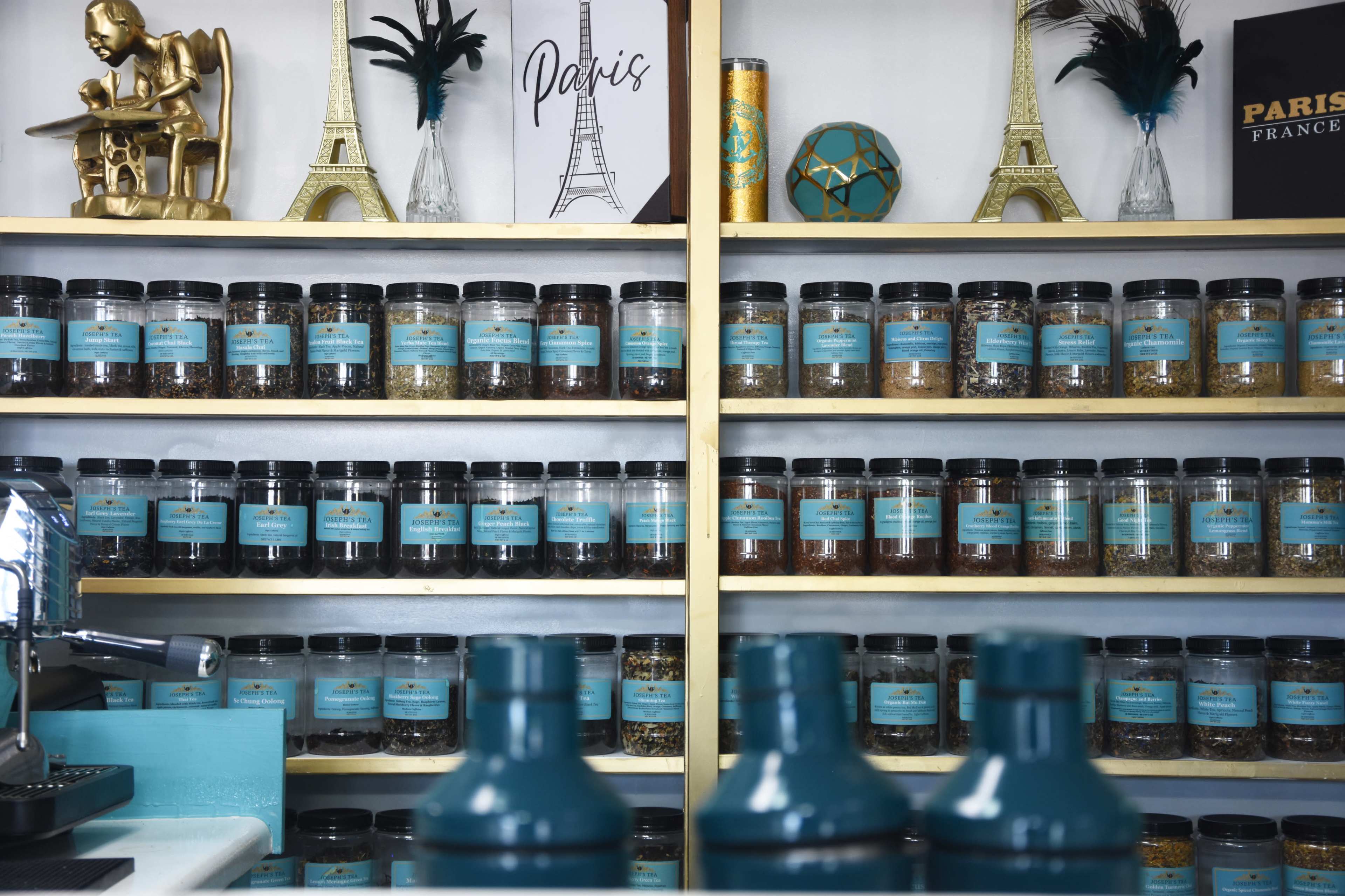 A display of jars filled with various loose teas arranged on shelves, with decorative Eiffel Tower items and a framed "Paris" sign in the background.